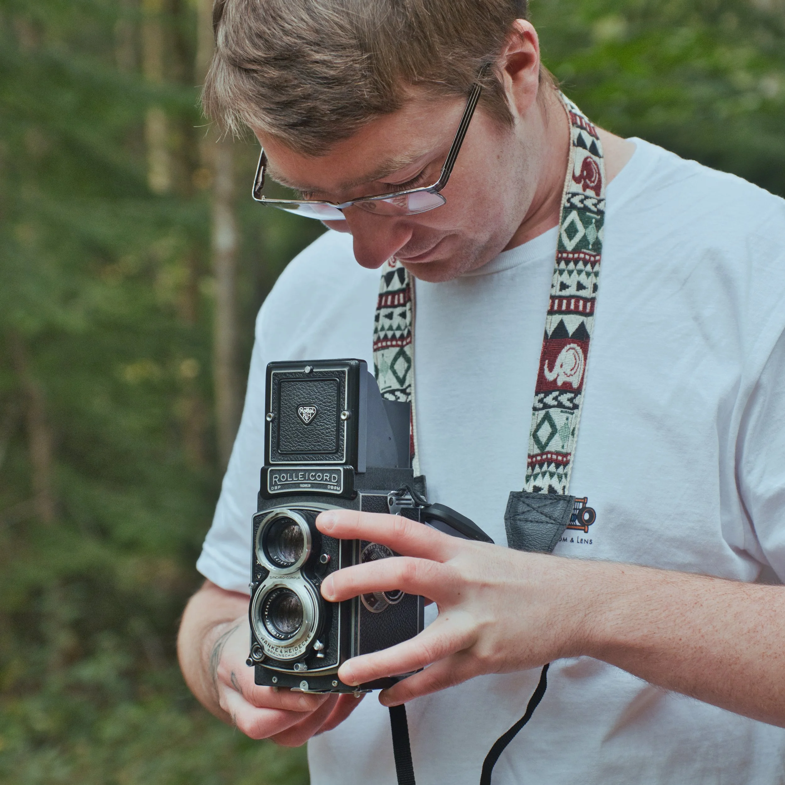 A man with glasses wearing a white t-shirt with a photography logo, holding a vintage twin-lens reflex camera outdoors with a wooded background.