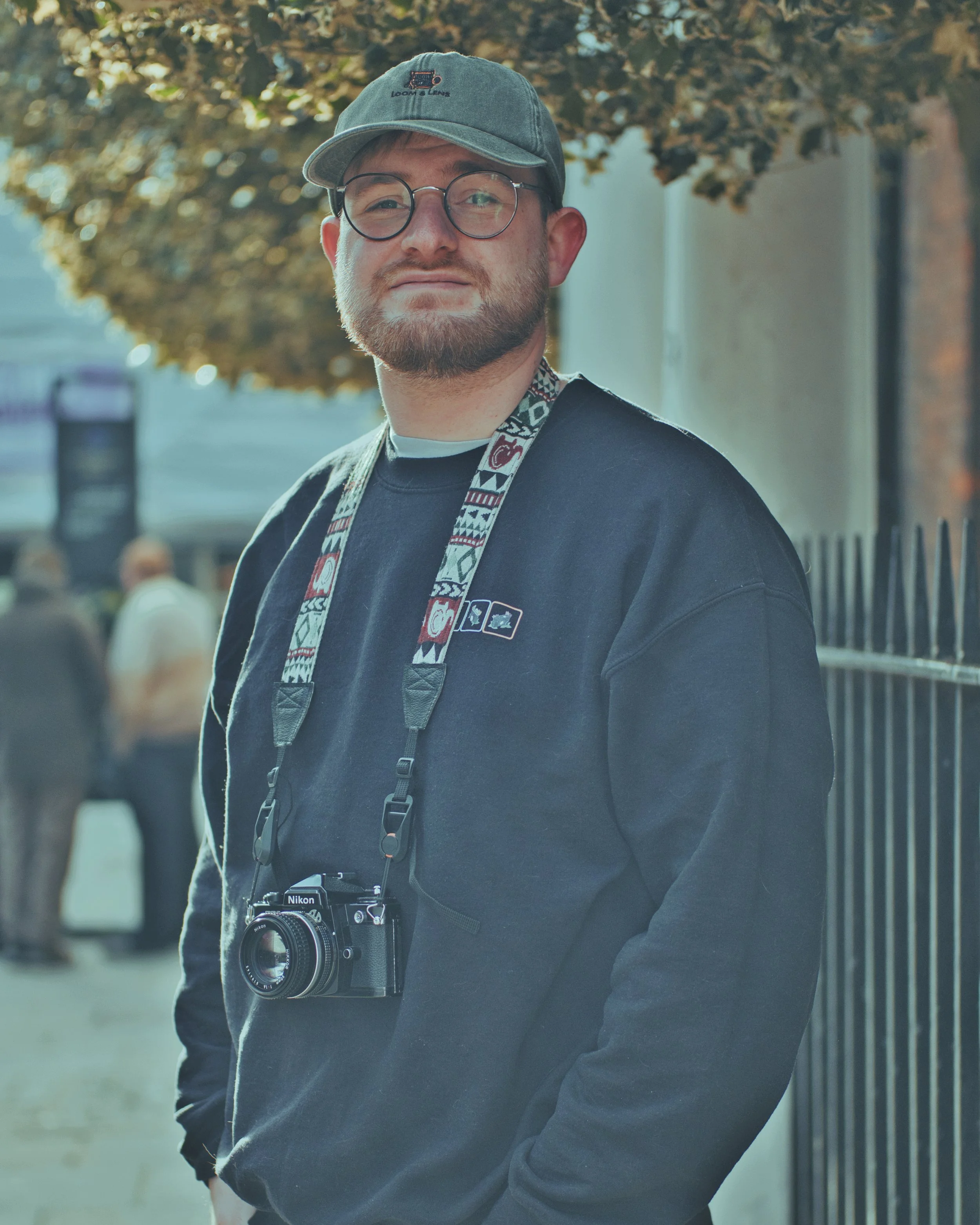 A man wearing glasses, a cap with a loom and lens logo, and a dark sweatshirt with a camera hanging around his neck, standing outdoors near a black metal fence and a tree with autumn-colored leaves.