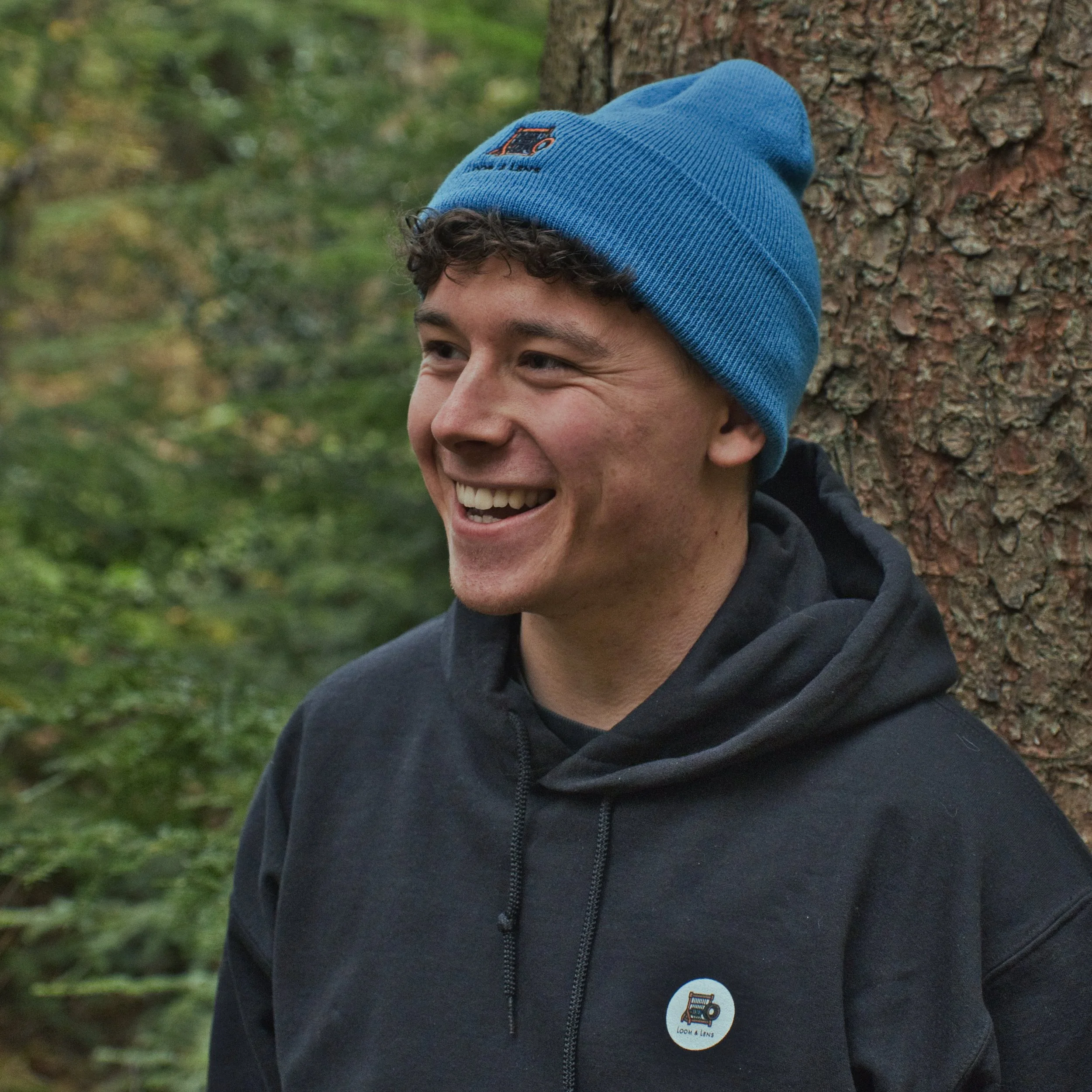 A young man smiling outdoors next to a tree, wearing a blue beanie and a black hoodie with a round badge.