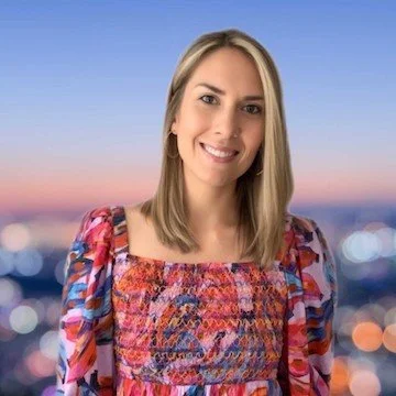 A young woman with blonde hair smiling, wearing a colorful patterned dress, standing outdoors during sunset with a blurred cityscape and bokeh lights in the background.