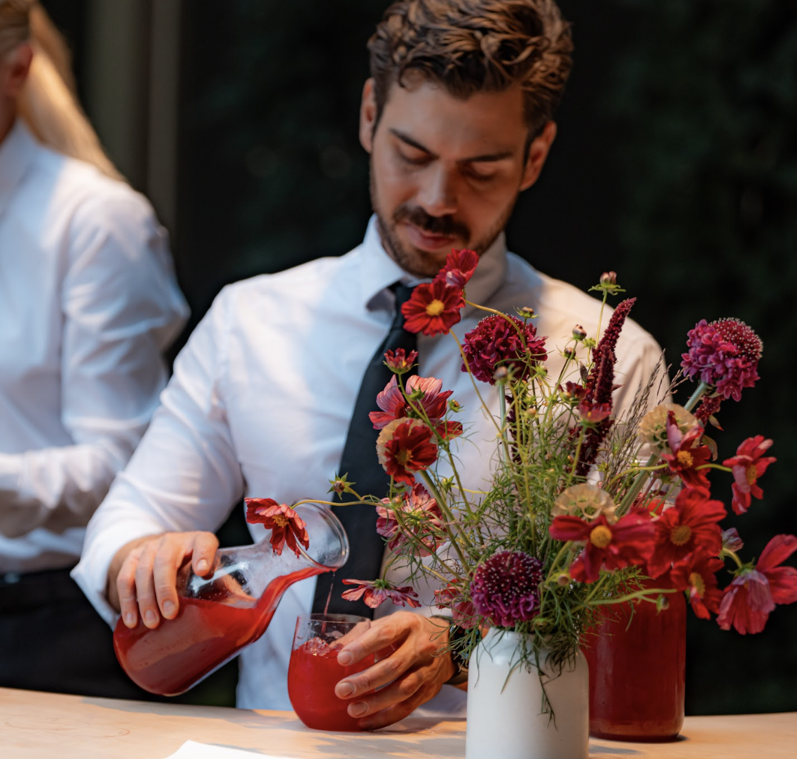 A man with dark hair and a beard in a white shirt and black tie pouring a red drink from a pitcher into a glass, with a bouquet of pink, red, and purple flowers in the foreground.
