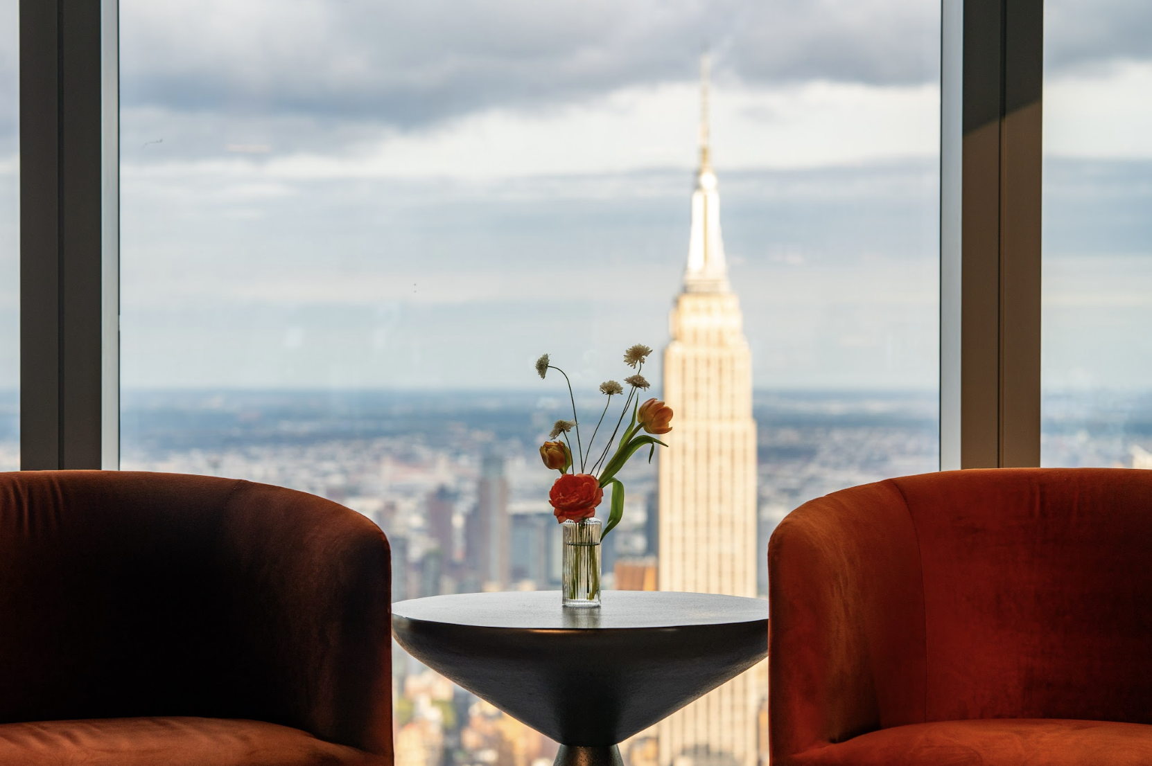 A view of the Empire State Building through a window from a cozy interior with a small round table, a glass vase with flowers, and two orange armchairs.