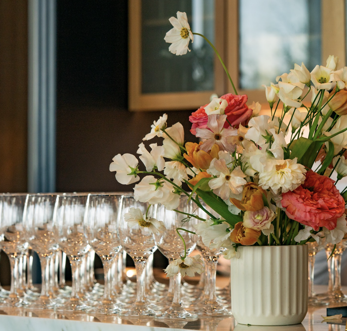 A floral arrangement in a white ribbed vase with white, pink, orange, and peach flowers on a table with empty wine glasses.