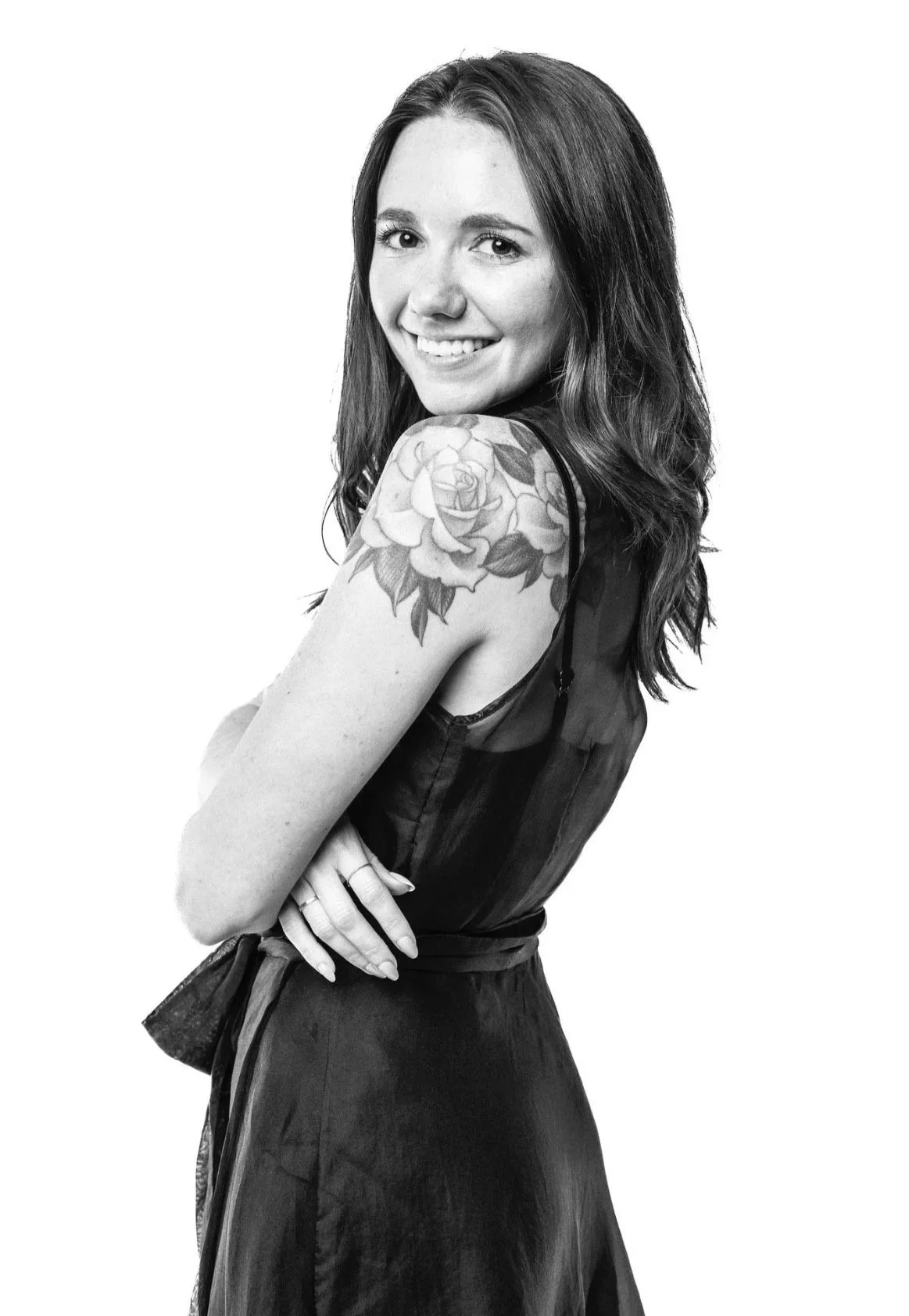A black and white photo of a smiling woman with long hair, wearing a sleeveless dress, showing a large floral tattoo on her shoulder.