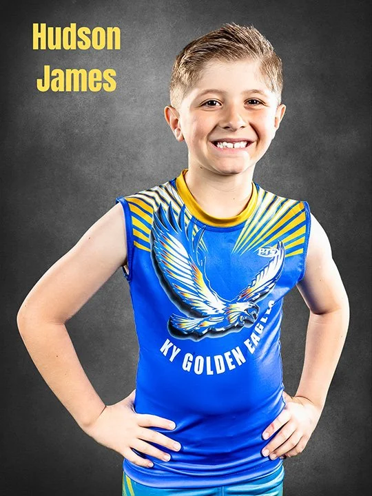Young boy smiling, wearing a blue sports jersey with a golden eagle and the text 'KY Golden Eagles' and 'Hudson James' on the upper left corner, against a dark gray background.