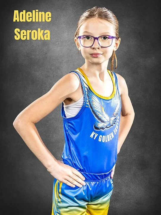 Young girl with glasses wearing a blue and yellow athletic uniform with a butterfly and the text "KY Golden Wings" standing with one hand on her hip against a dark background.