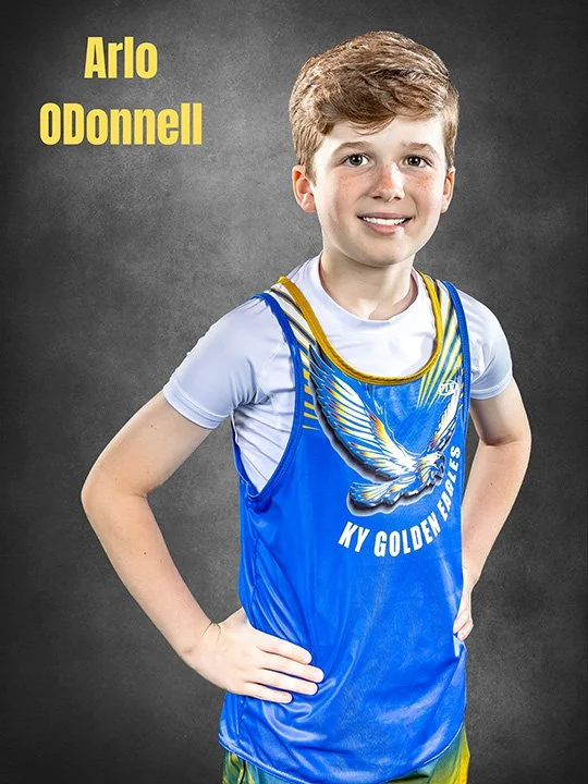 Young boy with brown hair wearing a blue track jersey with a design of an eagle and the words 'KY Golden Eagles' on it, standing with hands on hips and smiling against a dark gray background.