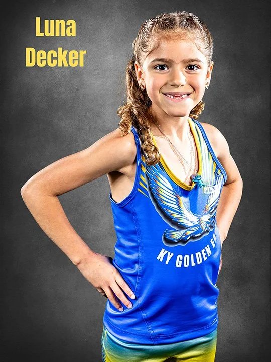 Young girl with curly hair wearing a blue athletic tank top with a bird graphic and the words "KY Golden E" on it, smiling with her hand on her hip against a gray background. Text in the top left corner says "Luna Decker."