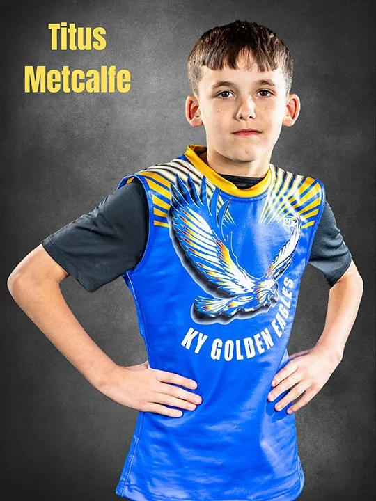 A young boy in a blue sports uniform with a large eagle graphic and the text 'KY Golden Eagles' stands with hands on hips, against a dark gray background. The name 'Titus Metcalfe' appears in yellow text in the upper left corner.