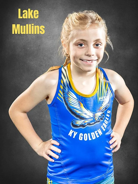 Young girl with blonde hair in a ponytail, wearing a blue athletic singlet with a winged eagle graphic and the words KY Golden Eagles, posing with hands on hips against a dark background.