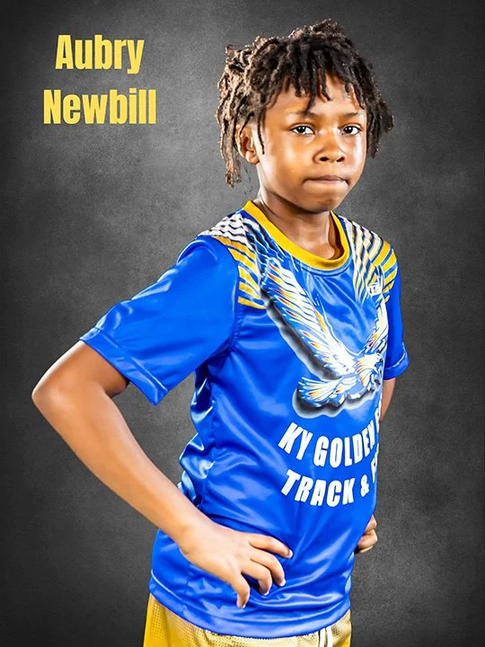 Young boy with dreadlocks wearing a blue sports jersey with a white eagle graphic and yellow accents, standing with hands on hips against a dark background, with names Aubry Newbill written in yellow text in the upper left corner.
