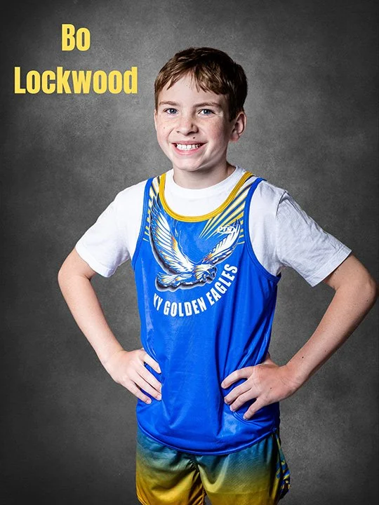 Young boy smiling, wearing a blue sports jersey with a golden eagle and the words "Golden Eagles," standing with hands on hips against a gray background.