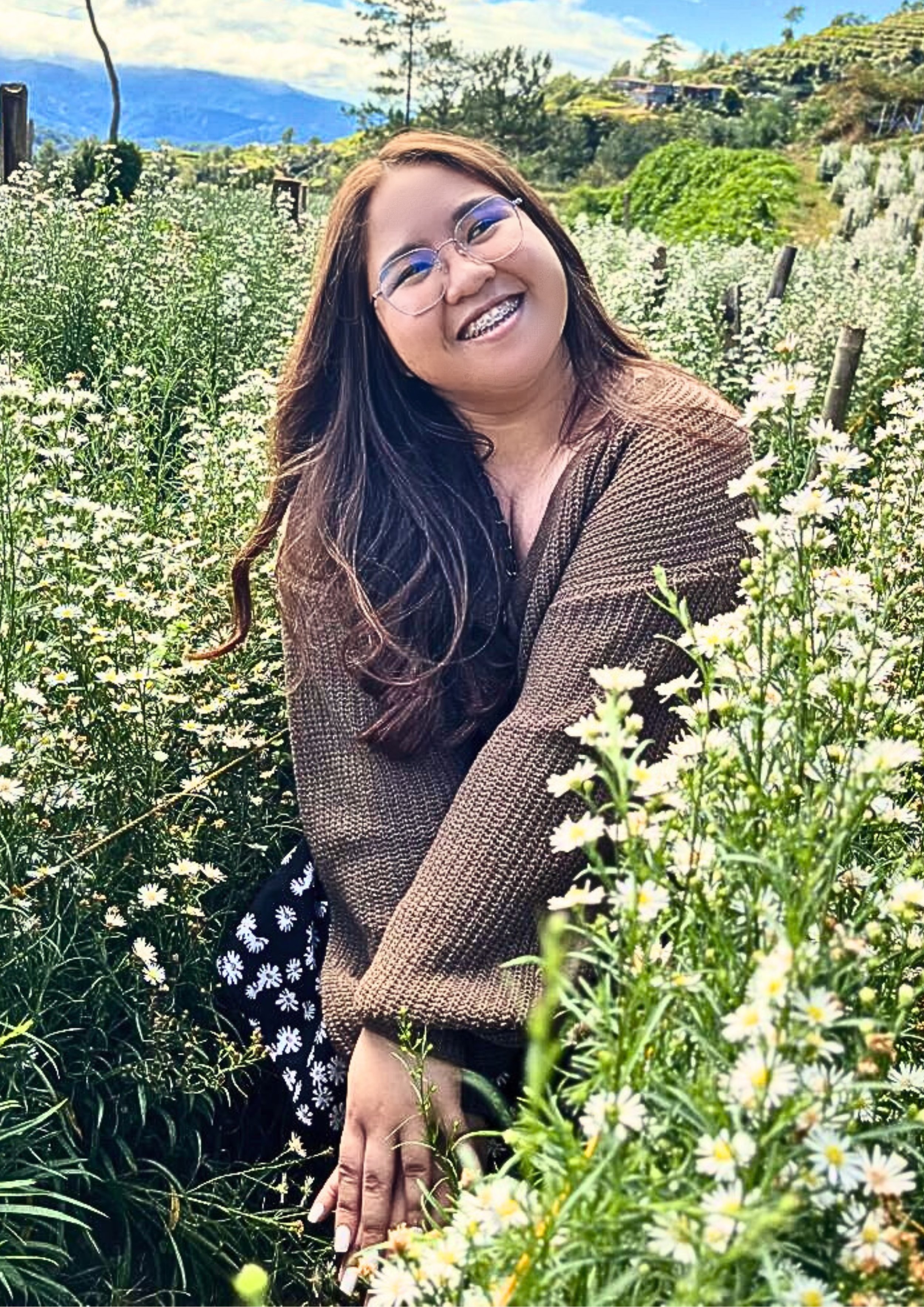 A woman with long dark hair and glasses smiling amid white daisies in a lush field with mountains and a cloudy sky in the background.