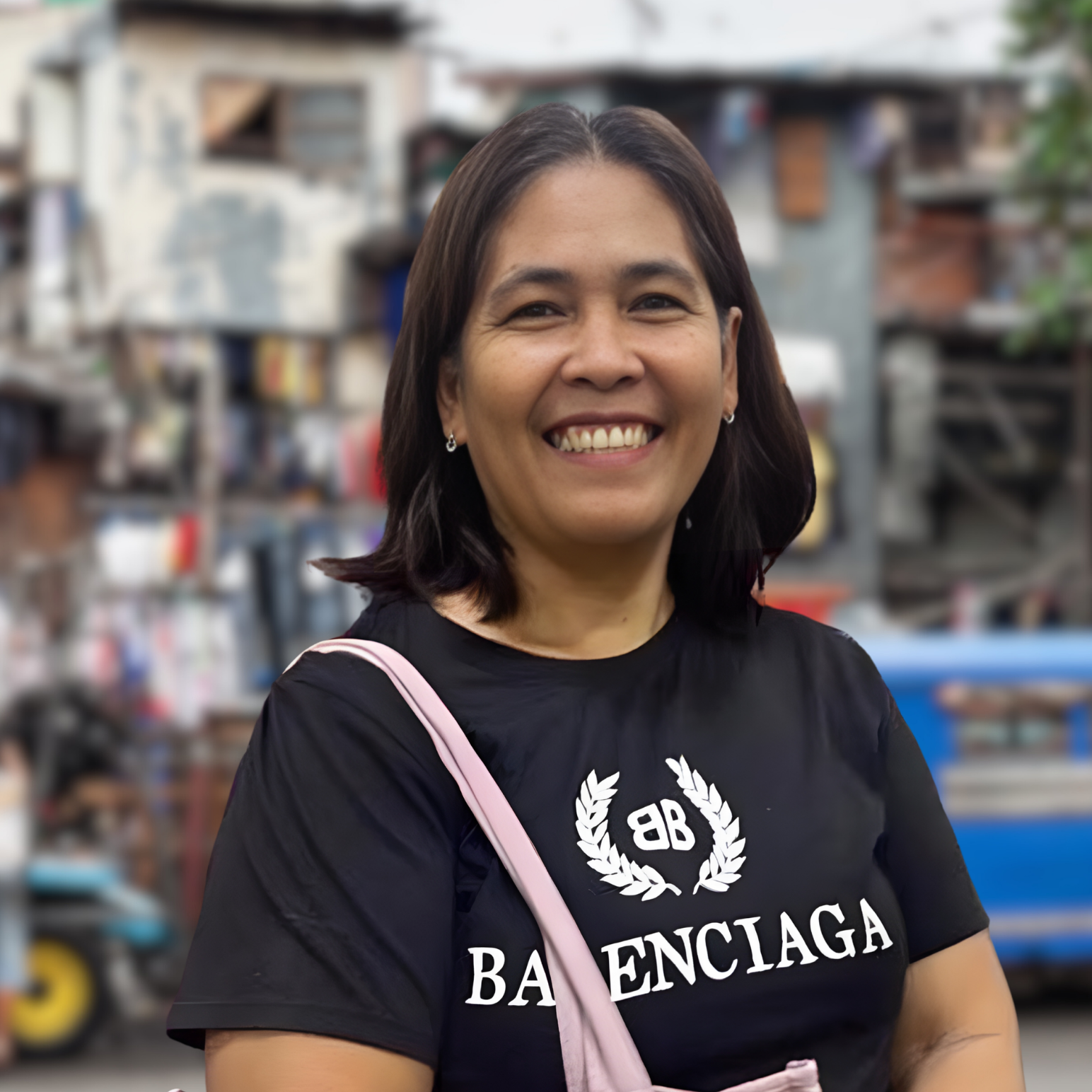 A smiling woman with shoulder-length dark hair, wearing a black Balenciaga t-shirt with white logo, standing outdoors in a background of a busy street scene.