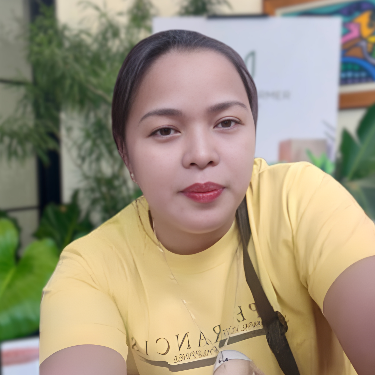 A young woman with short dark hair wearing a yellow shirt takes a selfie in an indoor setting with green plants in the background.