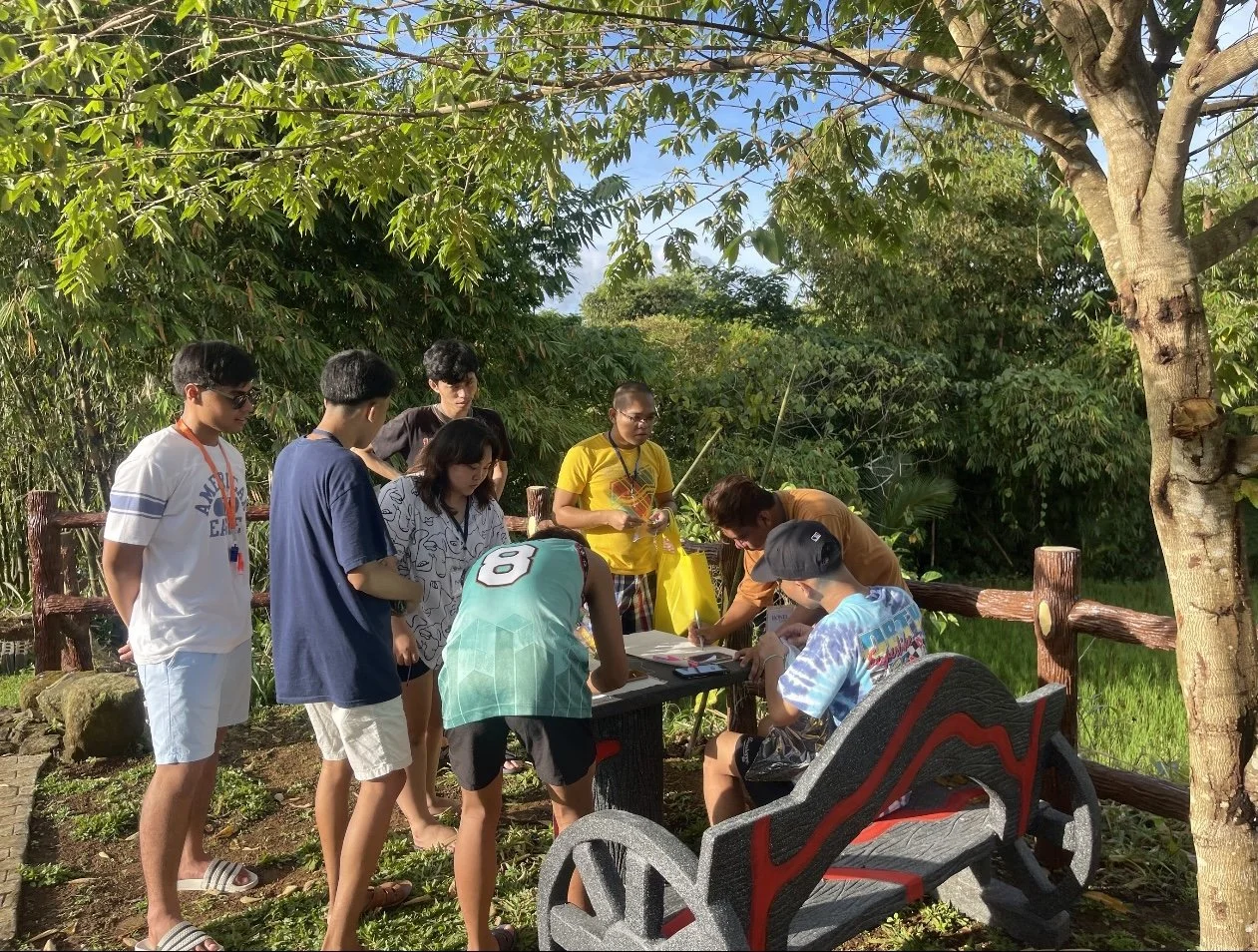 Group of young people gathered around a table outdoors in a green, wooded area with trees and bushes, some standing and some sitting, engaged in an activity, with sunlight filtering through the leaves.
