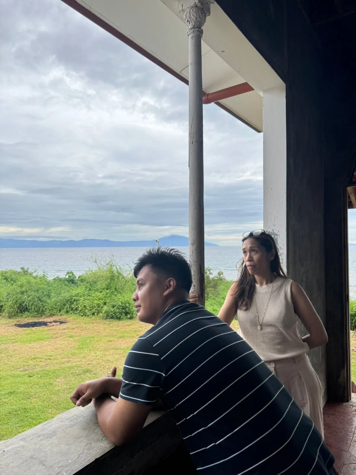 A man leaning on a railing and looking out at the ocean, with a woman standing behind him, on a covered porch overlooking a green landscape and the sea, with cloudy sky overhead.