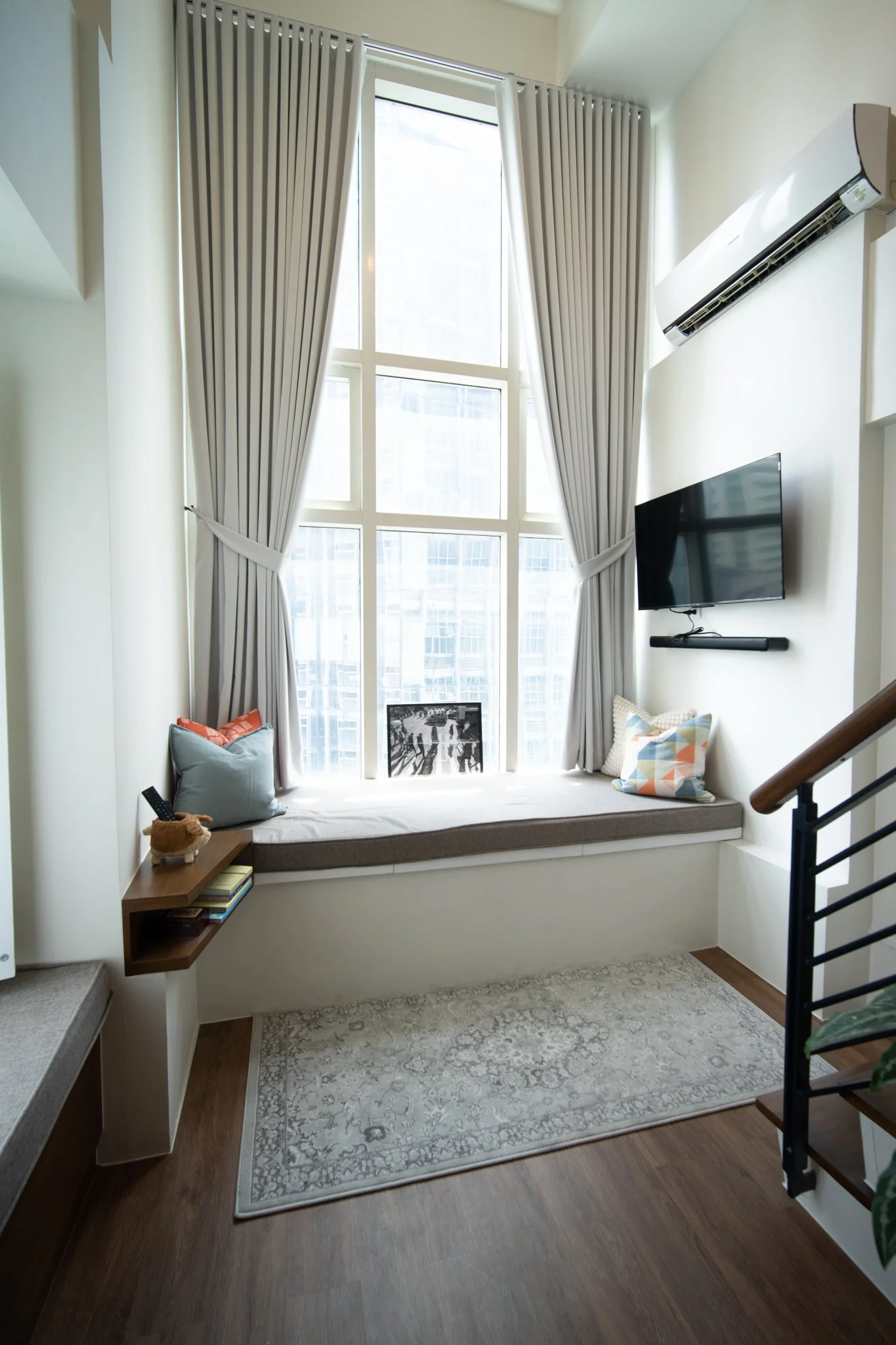 A cozy window nook with gray cushions, colorful pillows, an art piece, a small side table with books and a plush toy, positioned in front of large windows with gray curtains, an air conditioner unit, and a wall-mounted flat-screen TV in a modern interior.