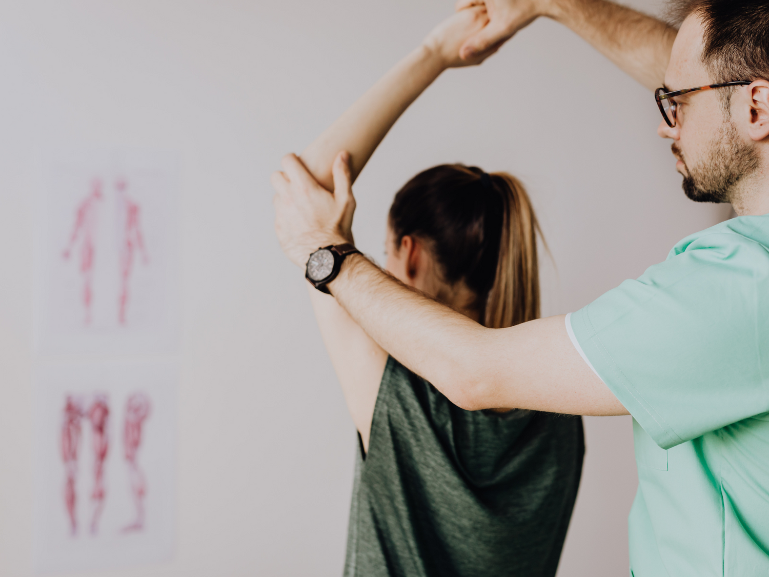 Massage therapist assisting a woman in stretching her arm overhead during a therapy session, with anatomical posters on the wall in the background.