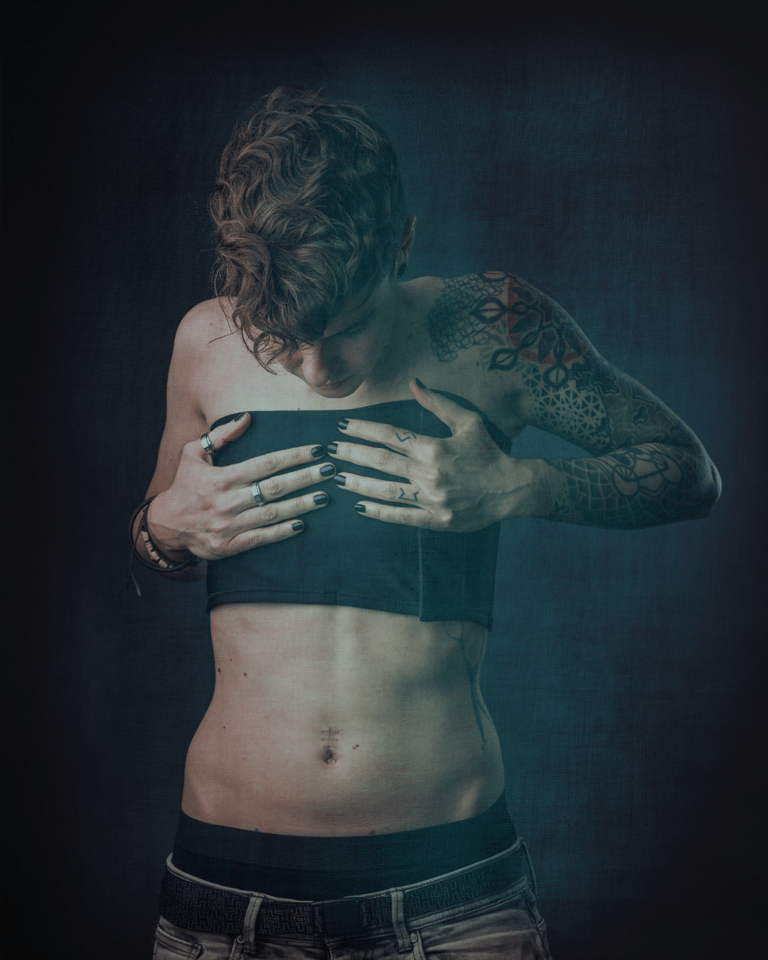A person with short curly hair, tattooed arms, and multiple rings on their fingers is wearing a chest binder and jeans, and looking at the appearance of the chest while wearing the binder.