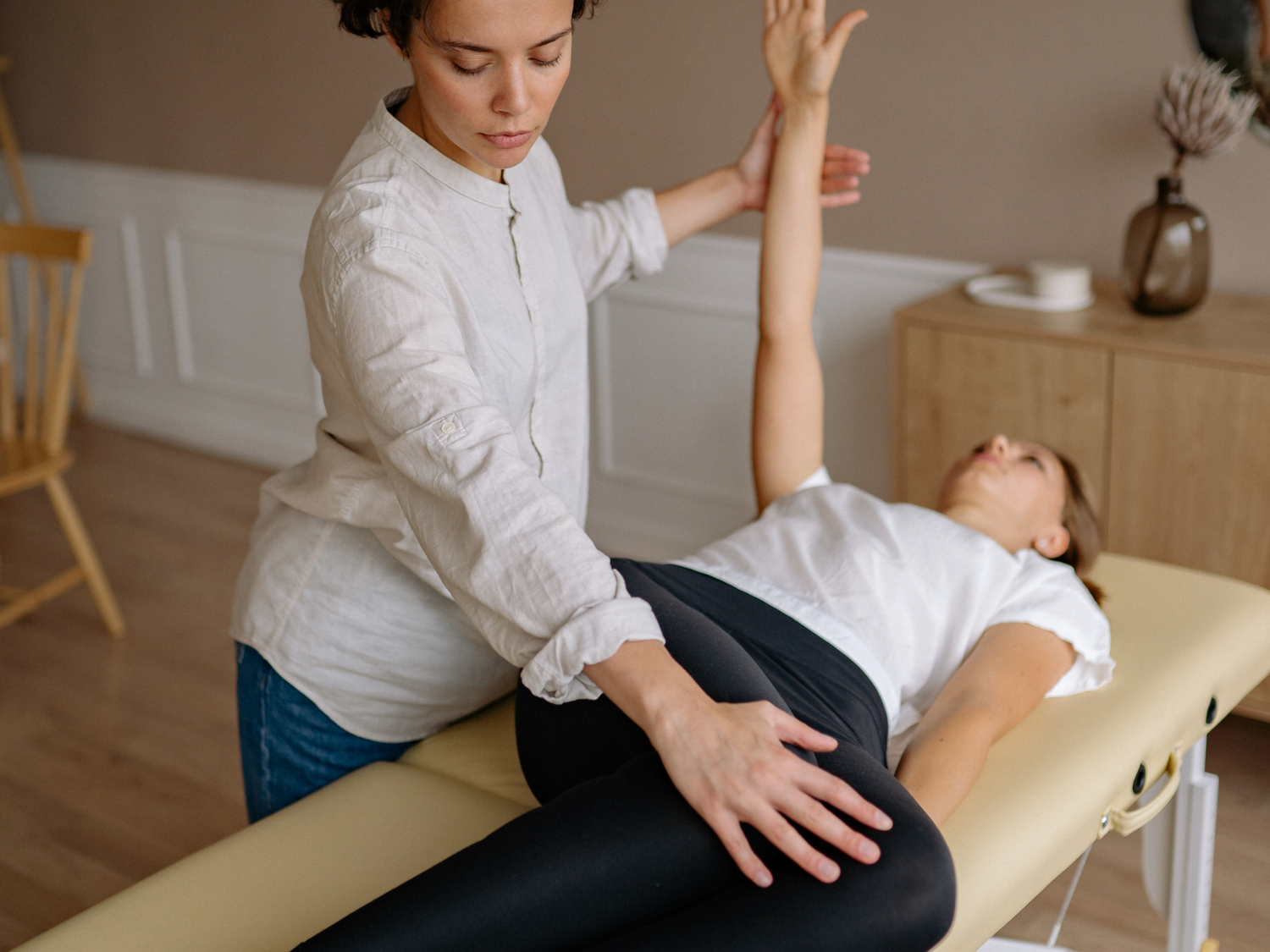 A woman lying on her side on a massage table with her arm raised, with a massage therapist providing a stretch, in a softly lit room with wooden furniture and a vase of flowers.