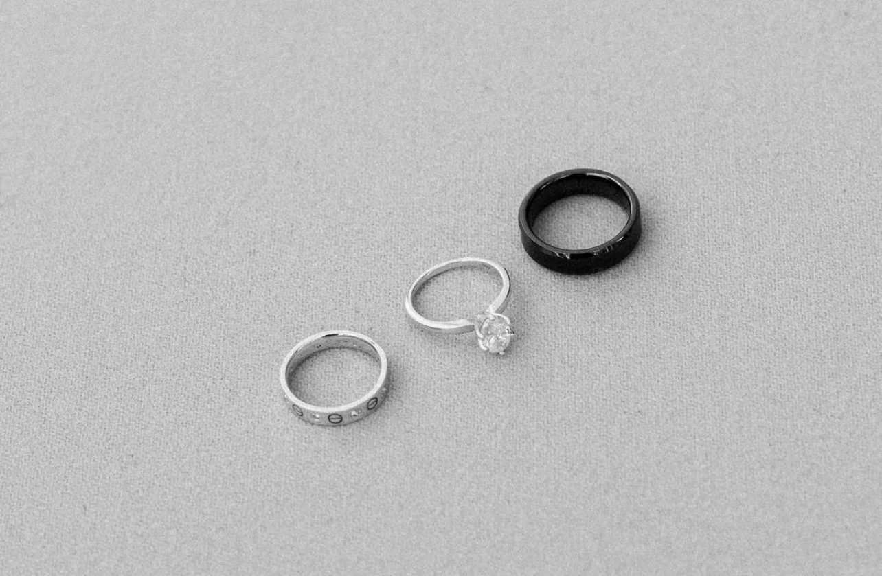 Three rings on a gray fabric surface: a black band, a silver band, and a silver ring with a diamond solitaire.