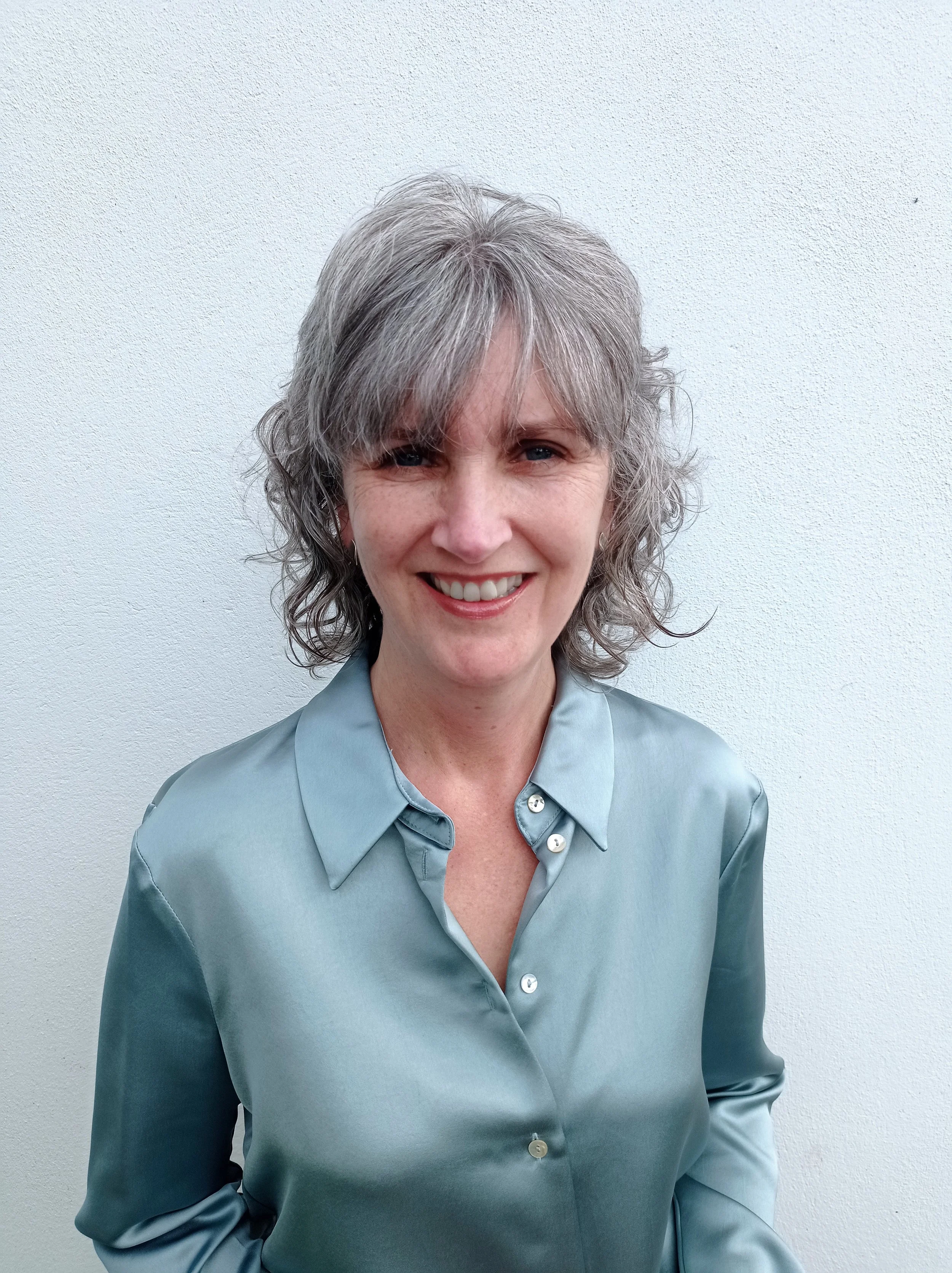 A woman with curly gray hair smiling, wearing a light blue satin blouse, standing against a plain white wall.