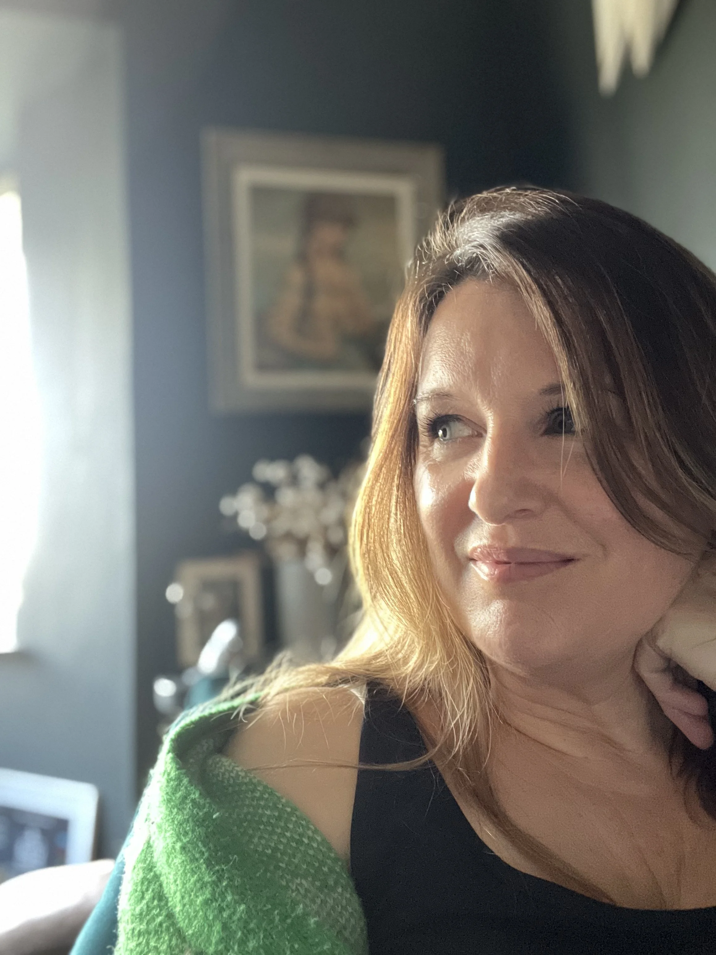 A woman with reddish-brown hair smiling softly, resting her chin on her hand in a brightly lit room with dark walls, framed artwork, and a window.