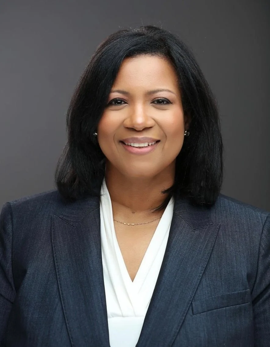 A professional woman with black hair, wearing a dark blazer and white blouse, smiling against a gray background.