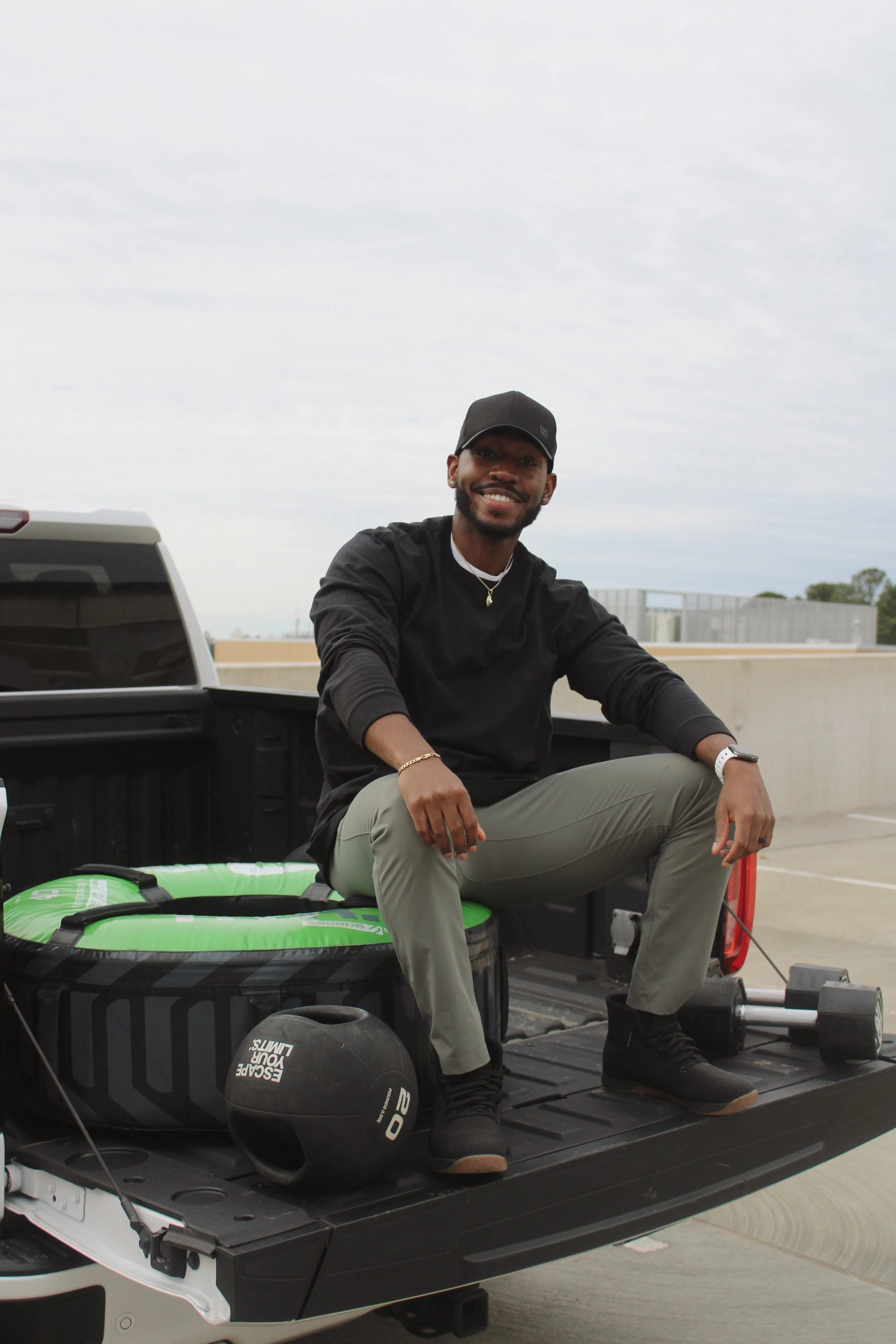 A smiling man in a black long-sleeve shirt, gray pants, and black shoes sitting on the tailgate of a pickup truck loaded with workout gear, including a green exercise mat, a black medicine ball, and an adjustable dumbbell, in a parking lot with an overcast sky.
