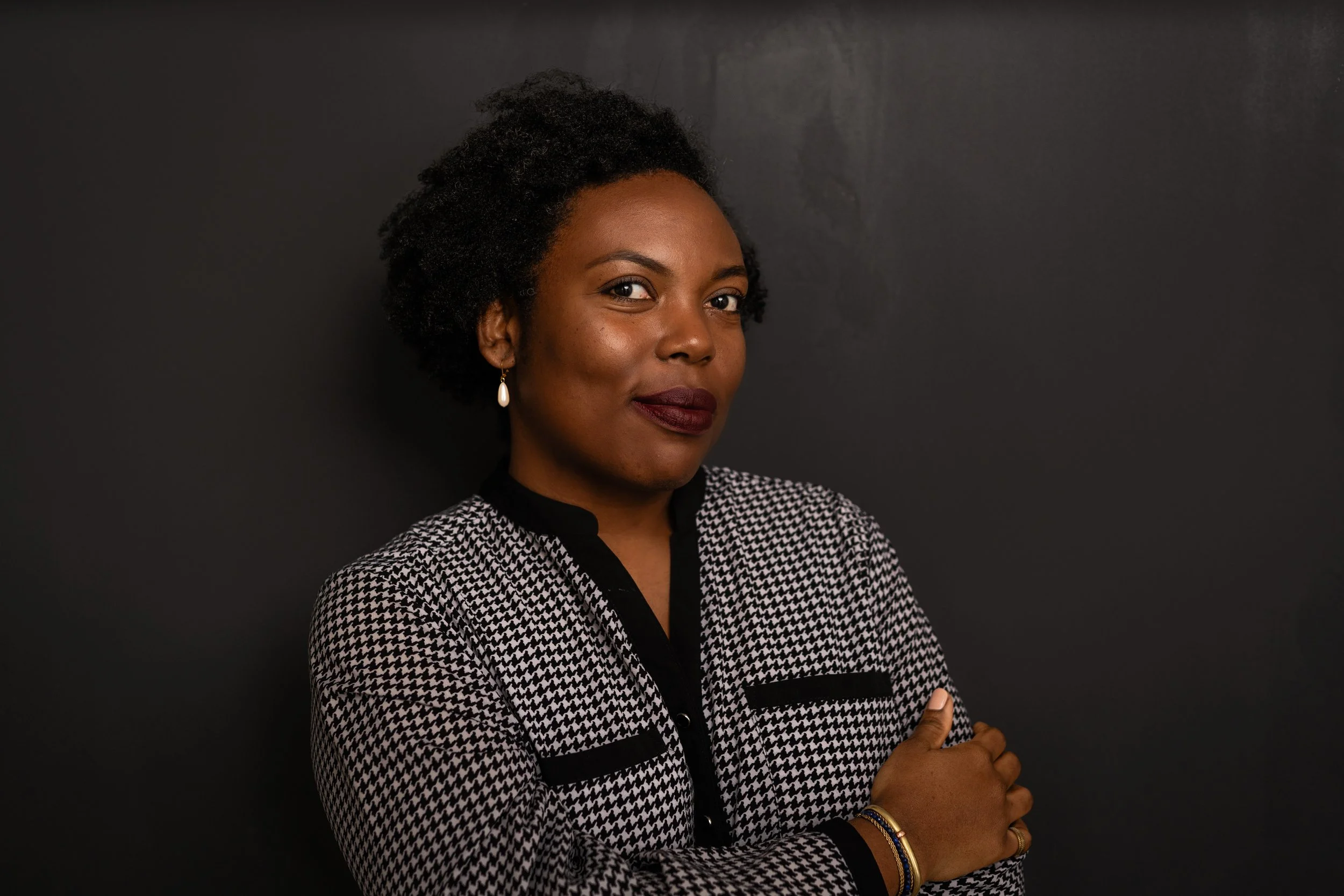 A confident African American woman with natural black hair, wearing a houndstooth patterned blouse, pearl earrings, and gold bracelets, standing against a dark background.