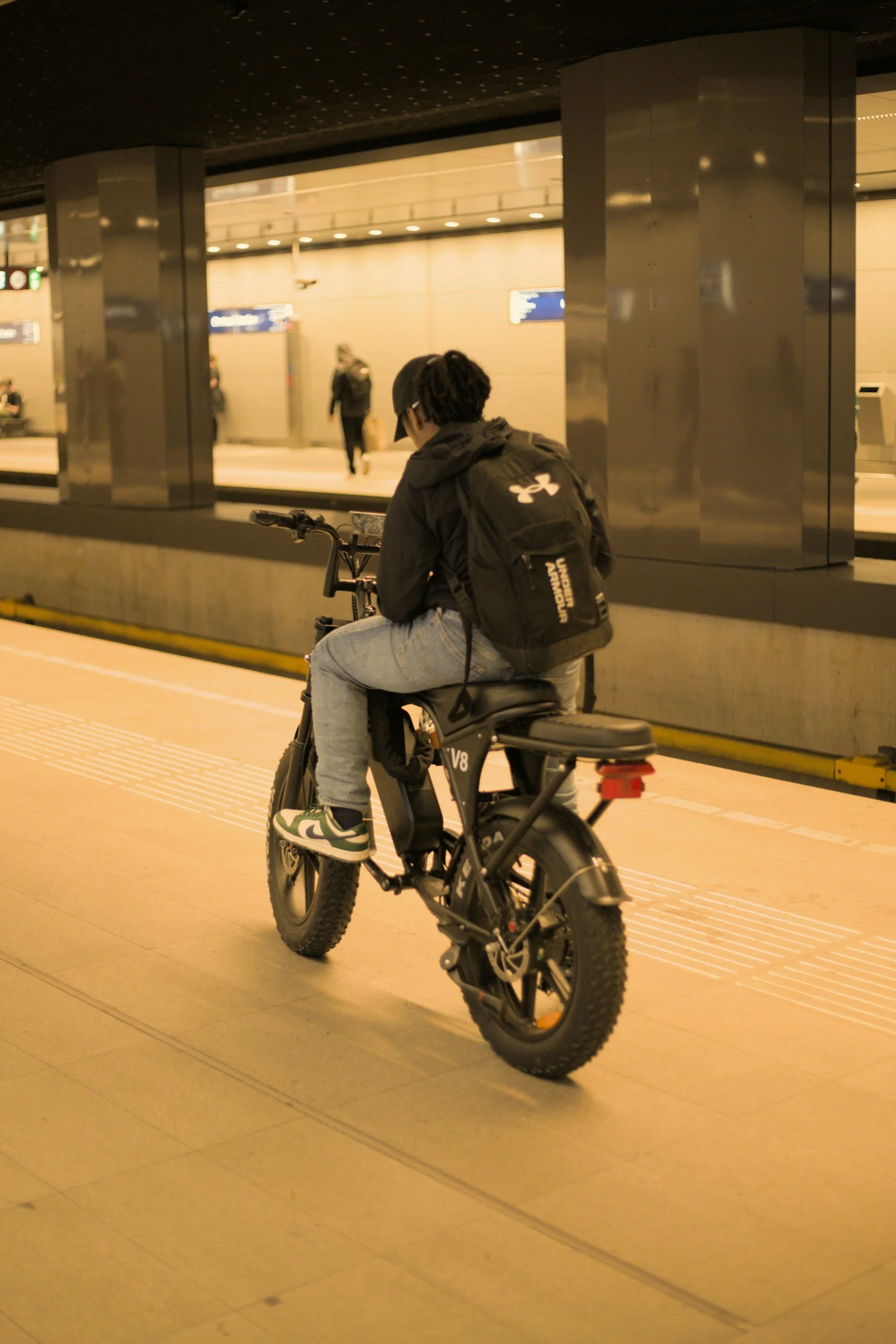 A person sitting on a black electric fatbike at a train station, with a backpack and wearing a black jacket and jeans, waiting near the platform as other travelers walk in the background.