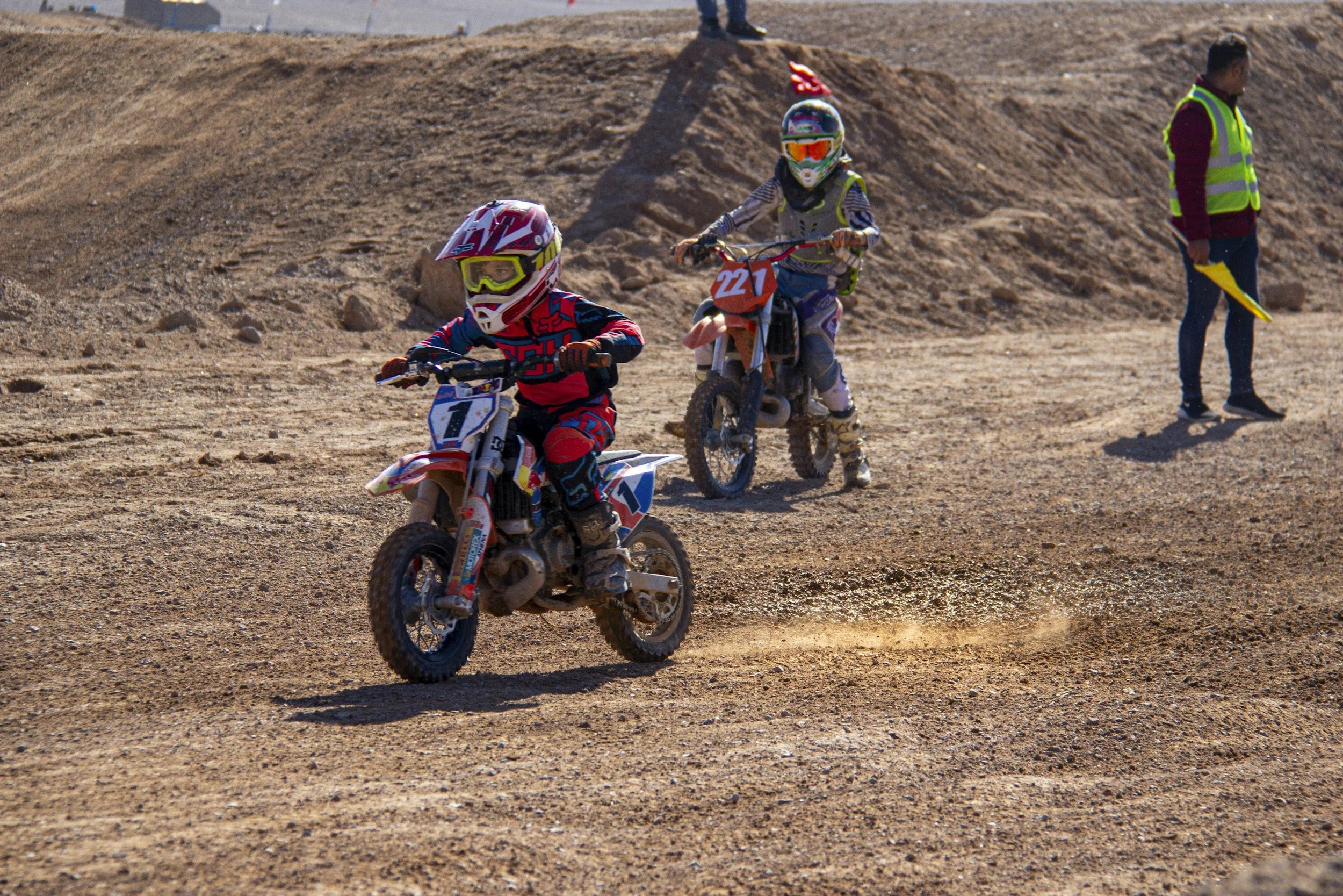 Two children riding dirt bikes on a dirt track, wearing helmets and protective gear, with a man standing nearby.