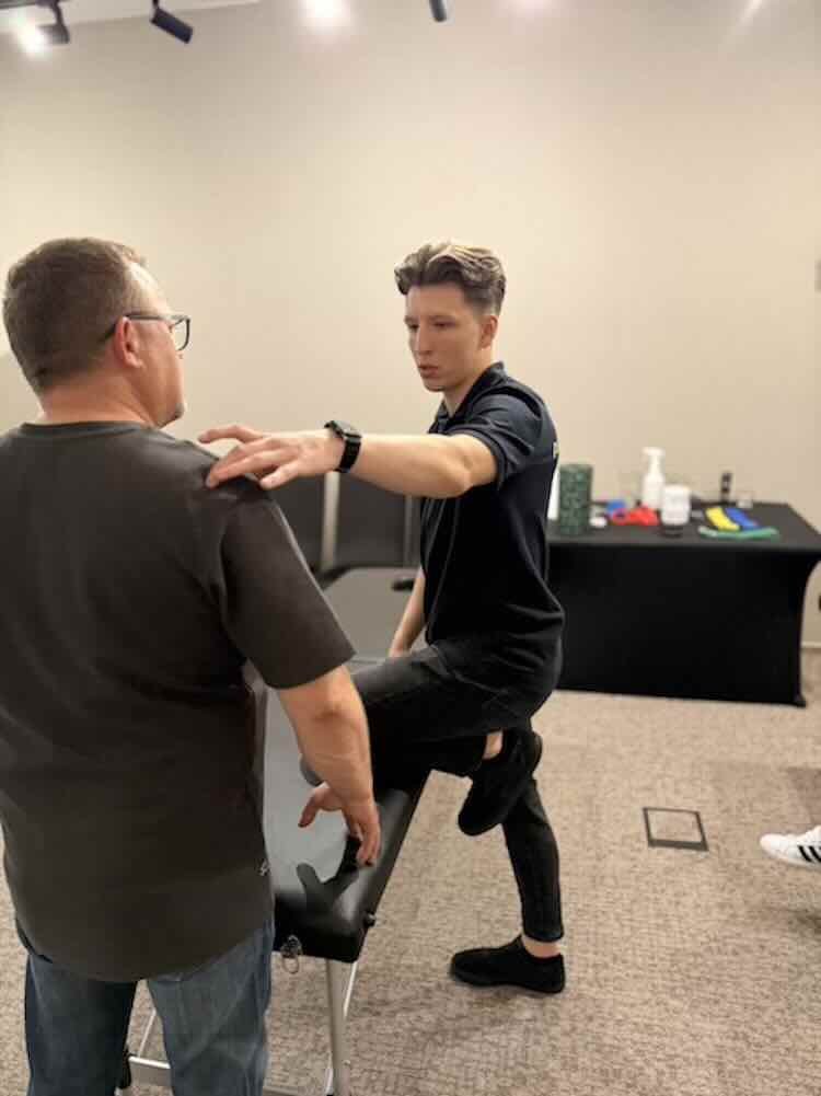 A young man is doing physical therapy or rehabilitation exercises with assistance from an older man in a clinical or therapy setting. The young man has his leg raised and rests on a padded table, while the older man supports his shoulder, and they appear to be engaged in a guided exercise or assessment.