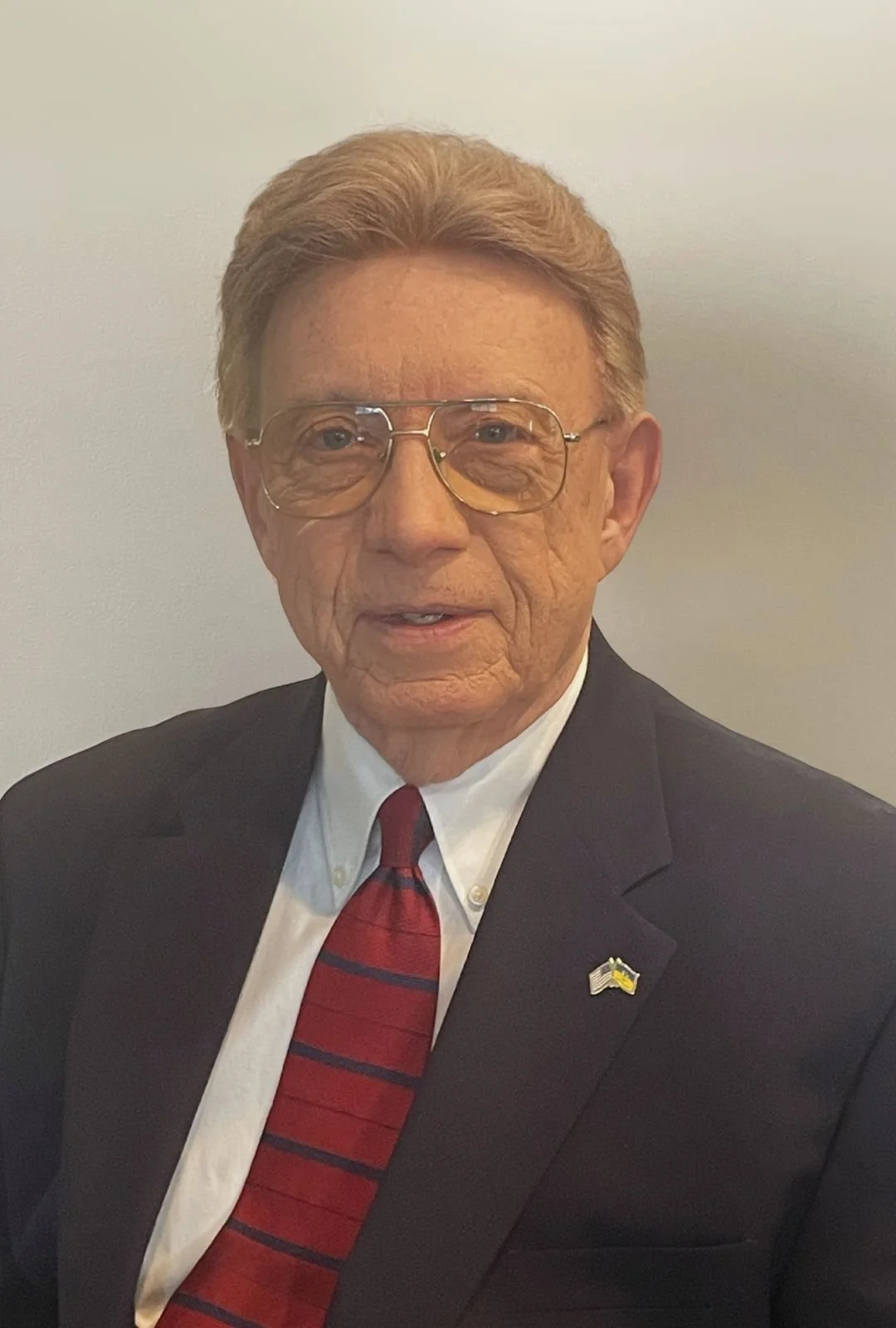 Gerry Tchir, wearing glasses, a formal suit with a white shirt, a red and black striped tie, and a small American flag pin on his jacket.