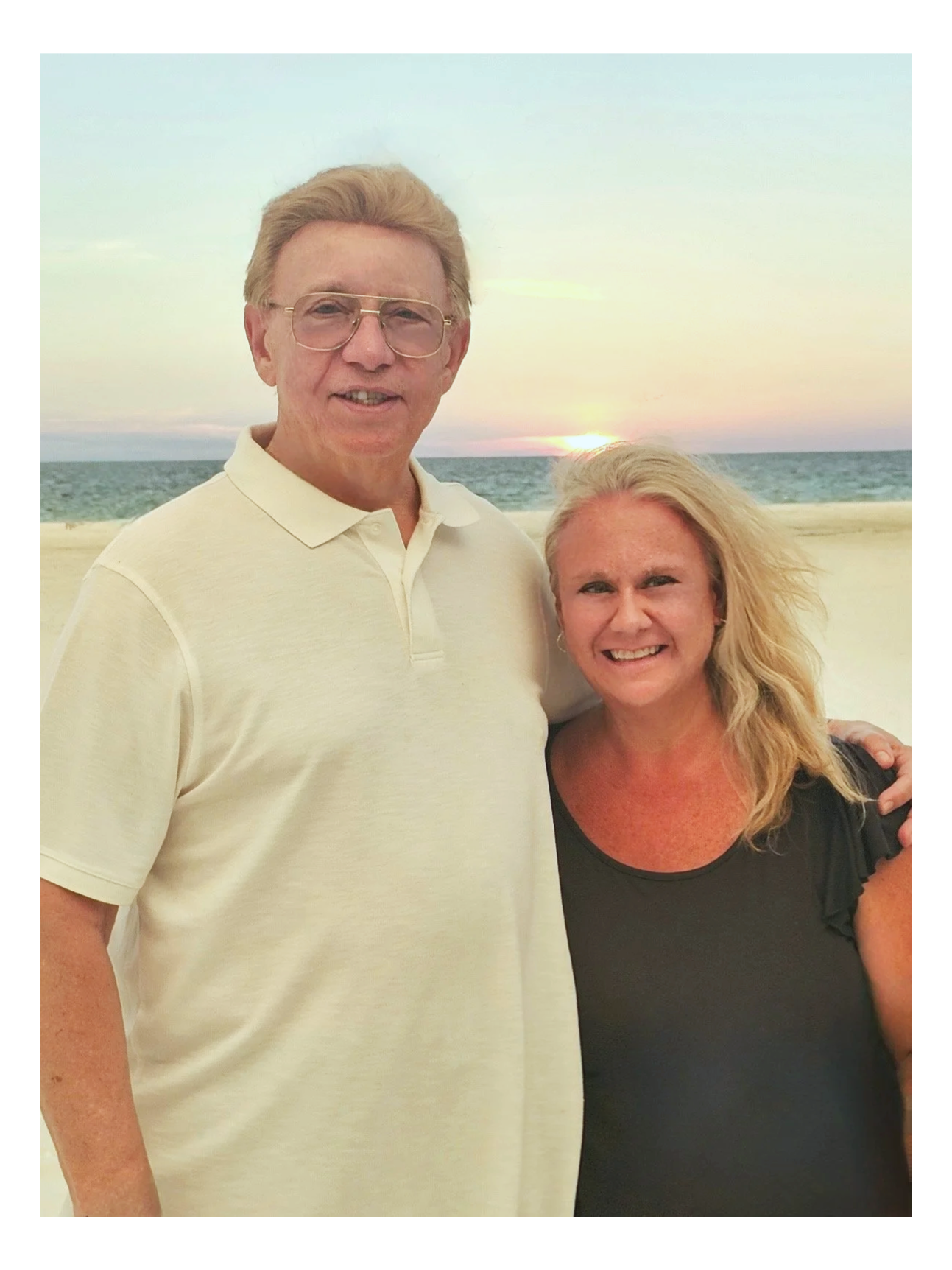 A man and woman standing on a beach at sunset, with the ocean and sky in the background. The man has blonde hair, glasses, and is wearing a light-colored polo shirt. The woman has blonde hair and is wearing a black top. They are smiling and posing for the photo.