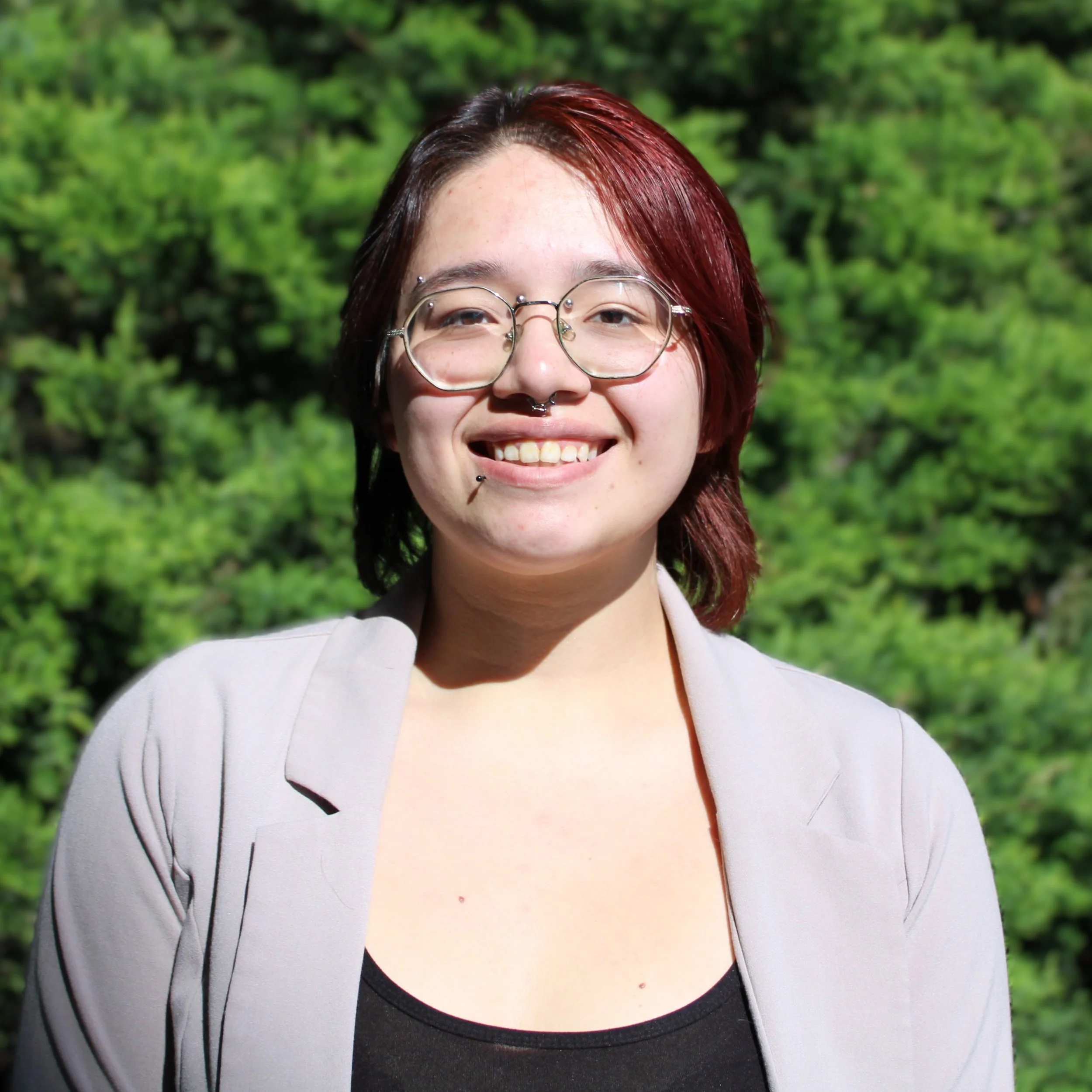 Person standing in front of bush, smiling, with short hair, glasses, white jacket,