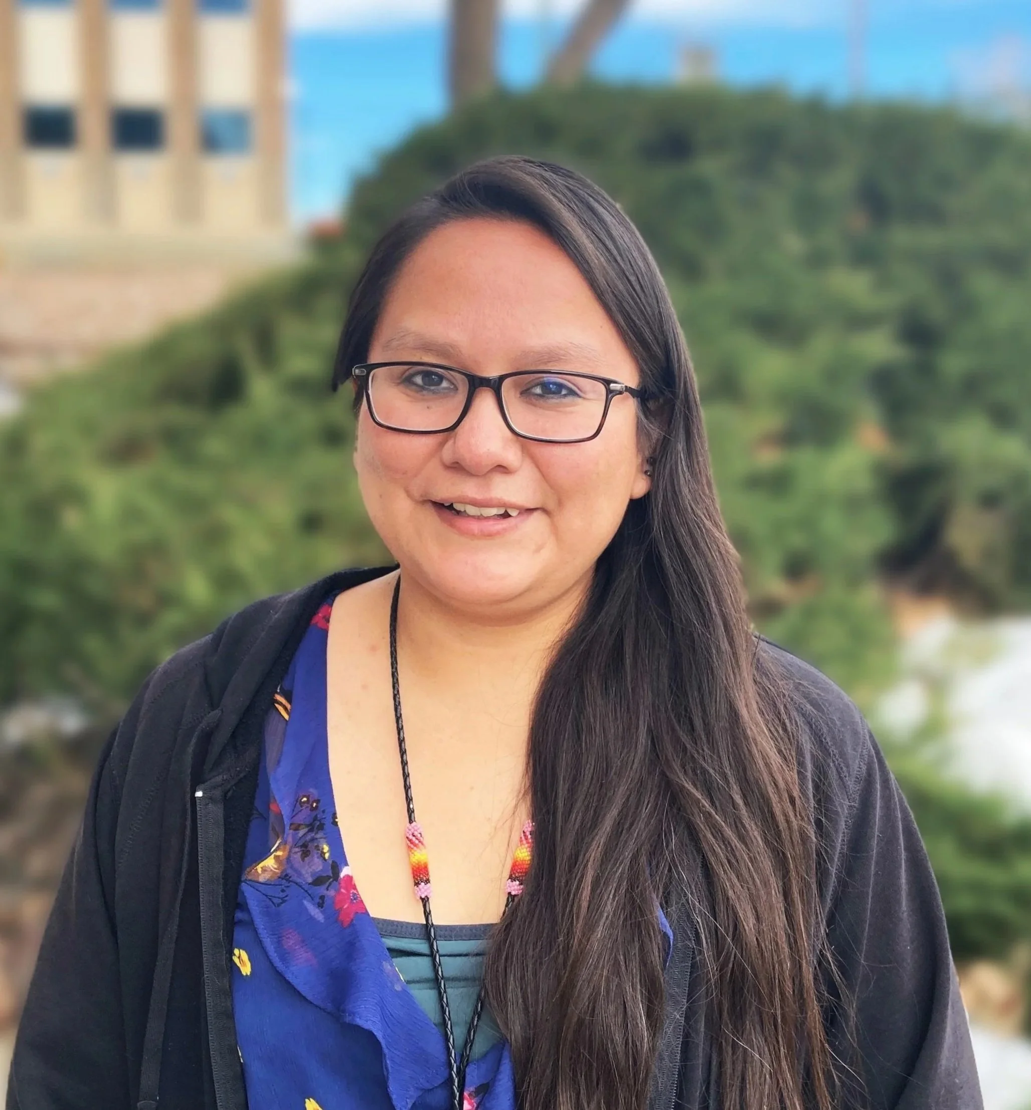 DIHFS Team - A young woman with glasses and long dark hair, standing outdoors in front of a blurred background of trees, a building, and water.