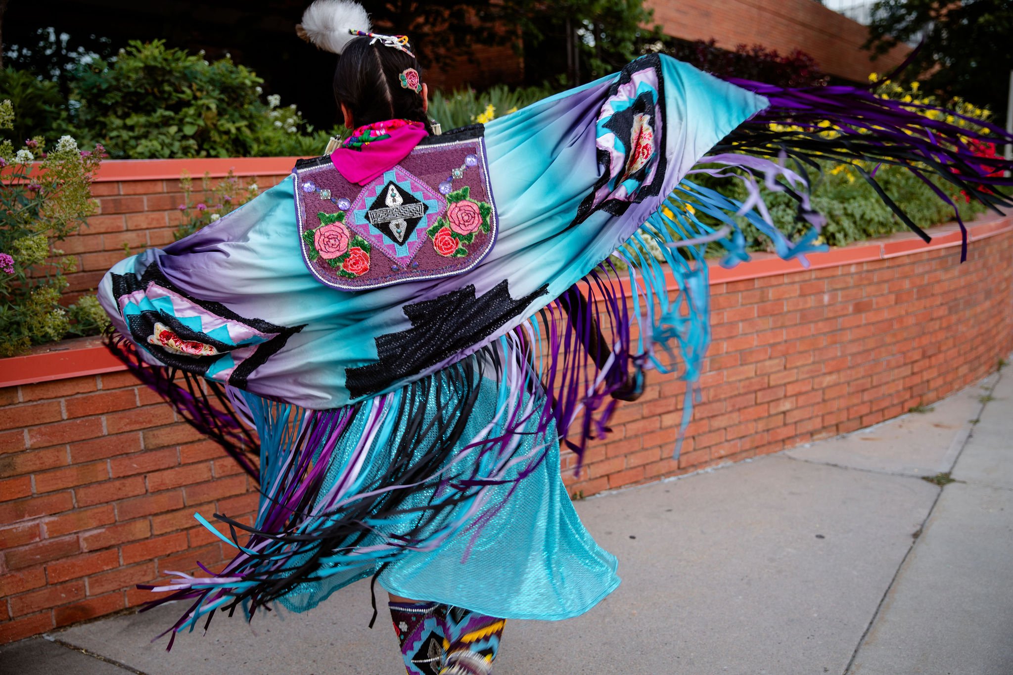 Person in colorful, fringed costume with intricate embroidery on back, walking past a brick wall and plants outdoors.