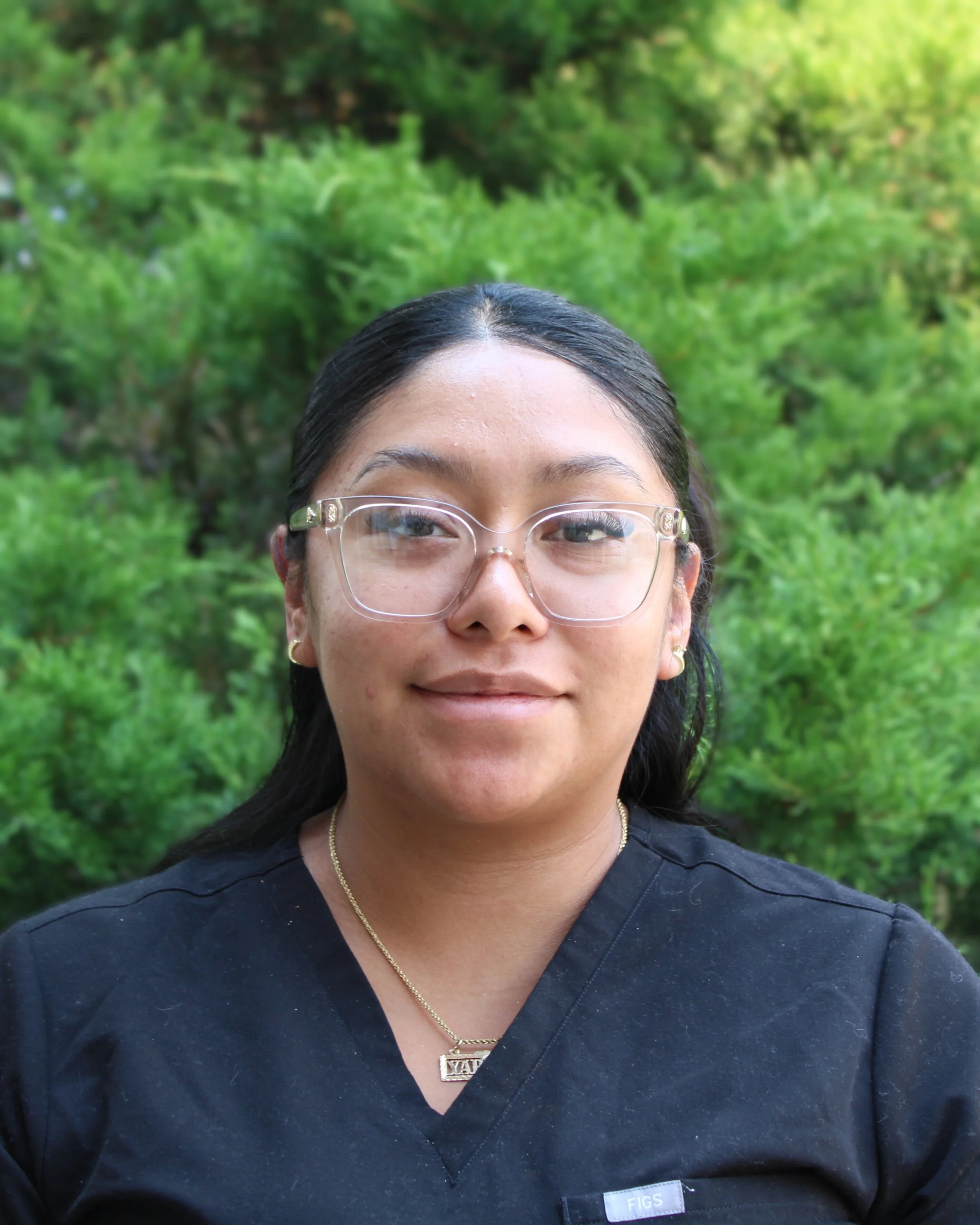 Woan with long dark hair, clear glasses, black scrubs, smiling
