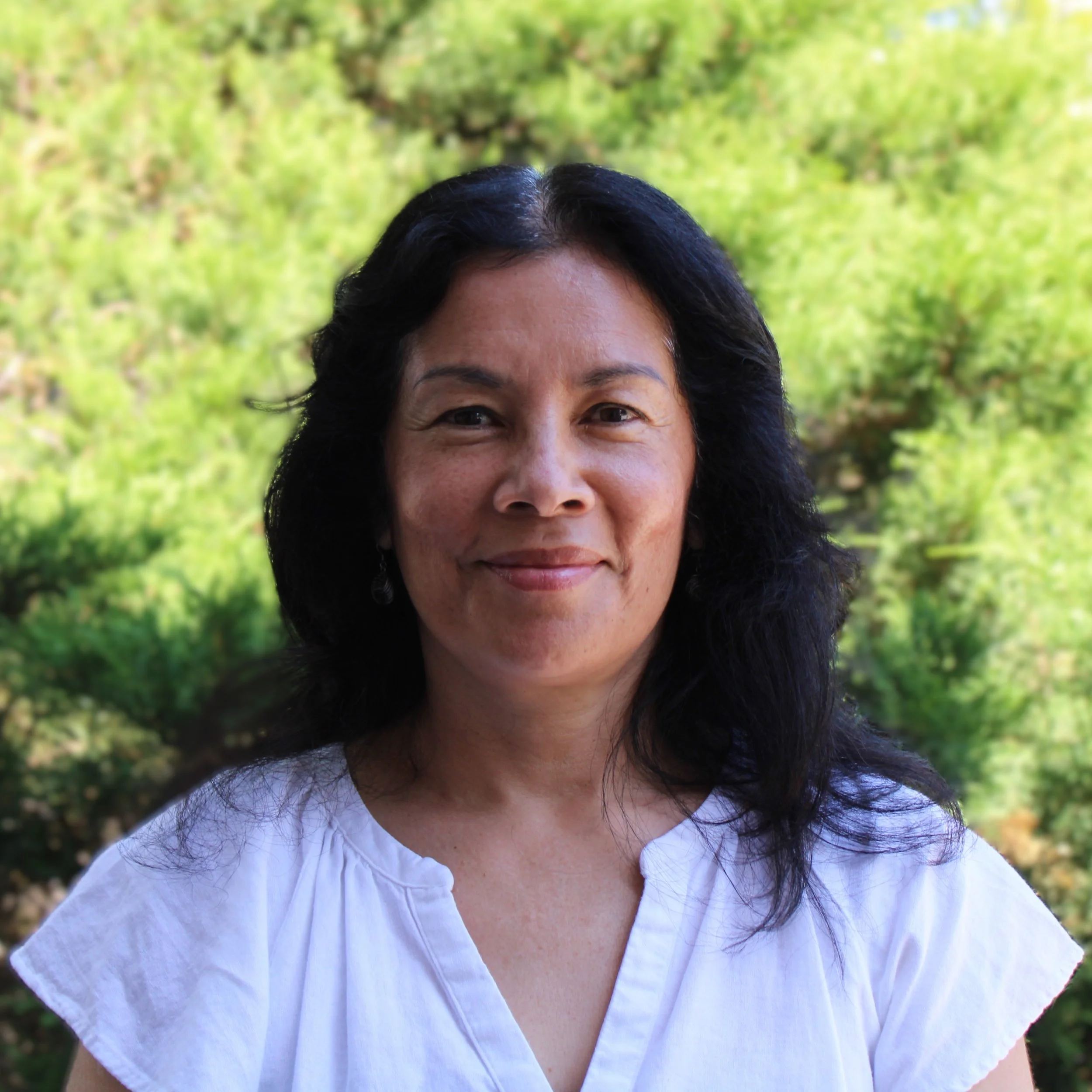 Woman with dark hair, white t-shirt, standing in front of bush, smiling, 