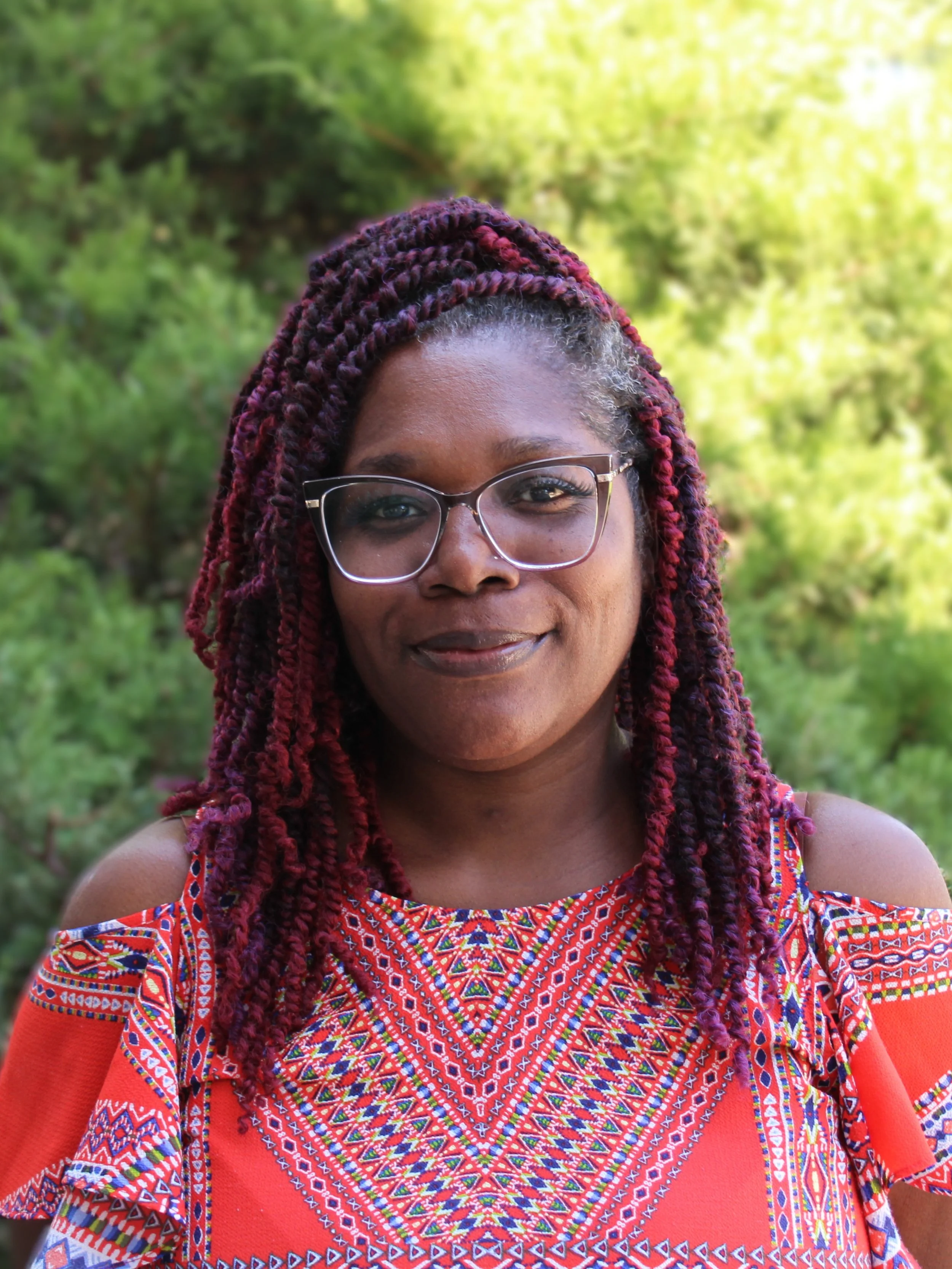 Woman with braided hair, red highlights, wearing a red patterned top, clear glasses, smiling, standing in front of bush