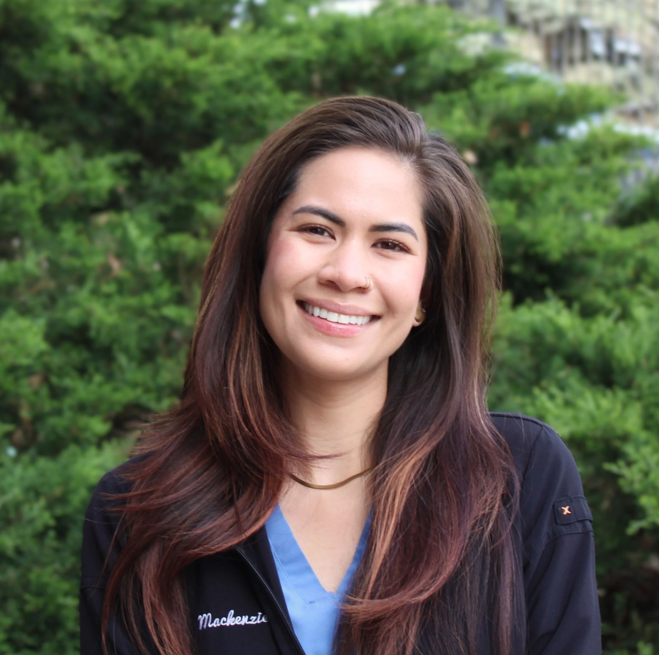 A woman with long dark brown hair wearing a black zip up jacket over a blue v-neck shirt smiles at the camera in front of a green shrub background