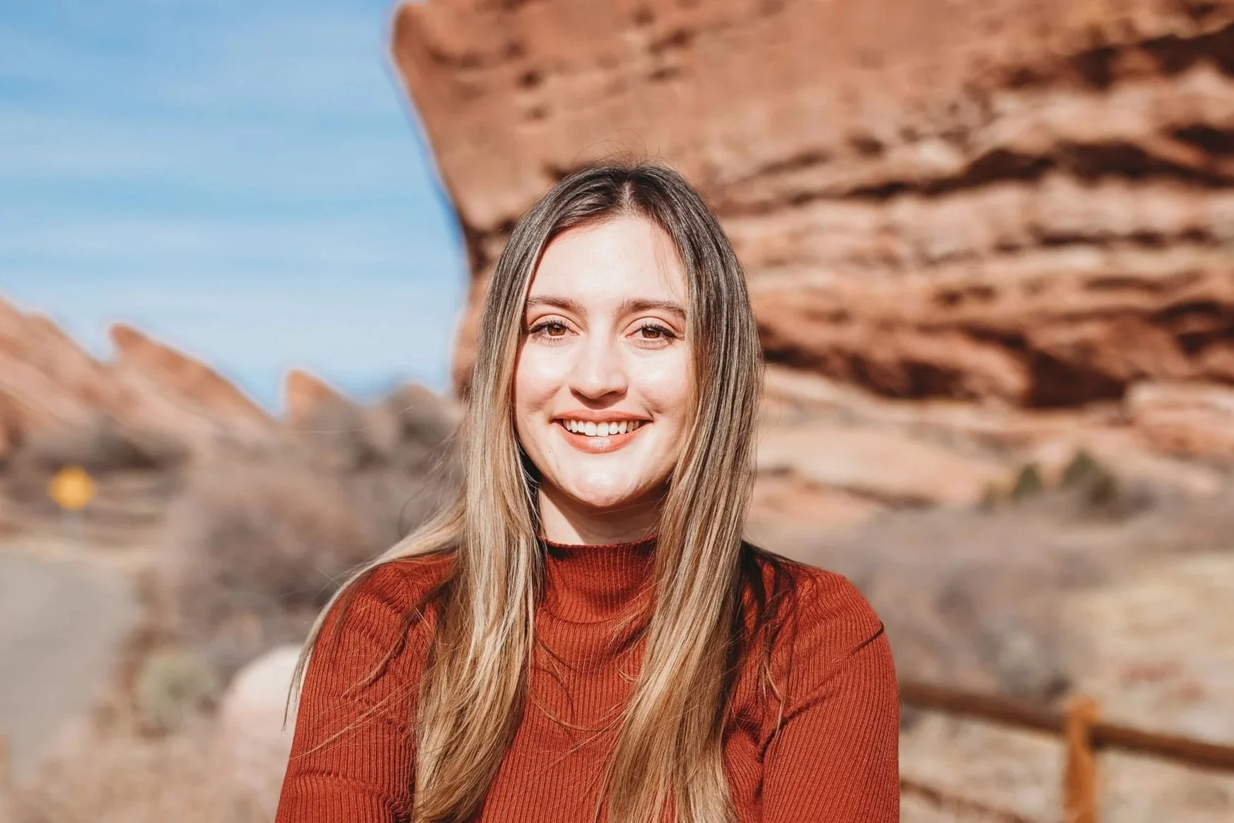 A young woman with long blonde hair smiling outdoors in a desert landscape with red rock formations and a wooden fence.