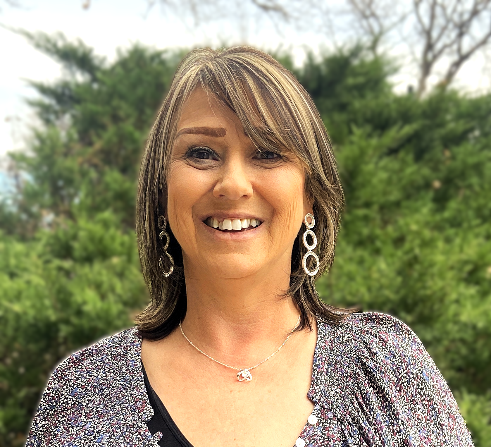 Woman with sideswept bangs and shoulder length hair smiles in front of a background of greenery