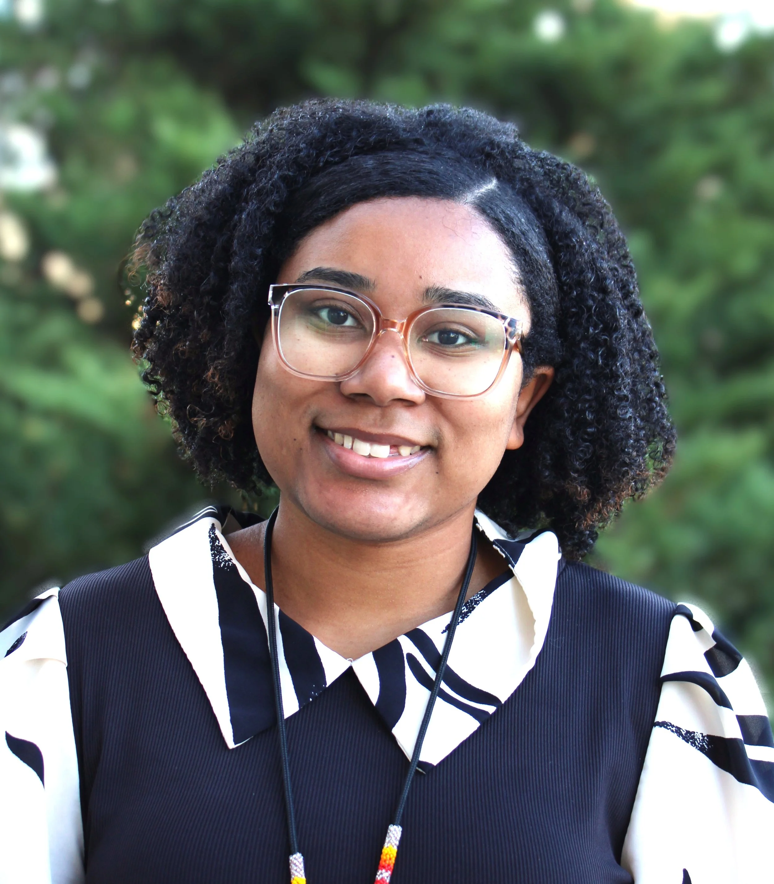 DIHFS Team - A woman smiling outdoors with green trees in the background, wearing a green top layered with a tan fleece jacket, and silver hoop earrings.