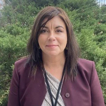 DIHFS Team - A female medical provider standing outdoors in front of a green shrub, wearing a deep purple jacket, smiling at the camera.