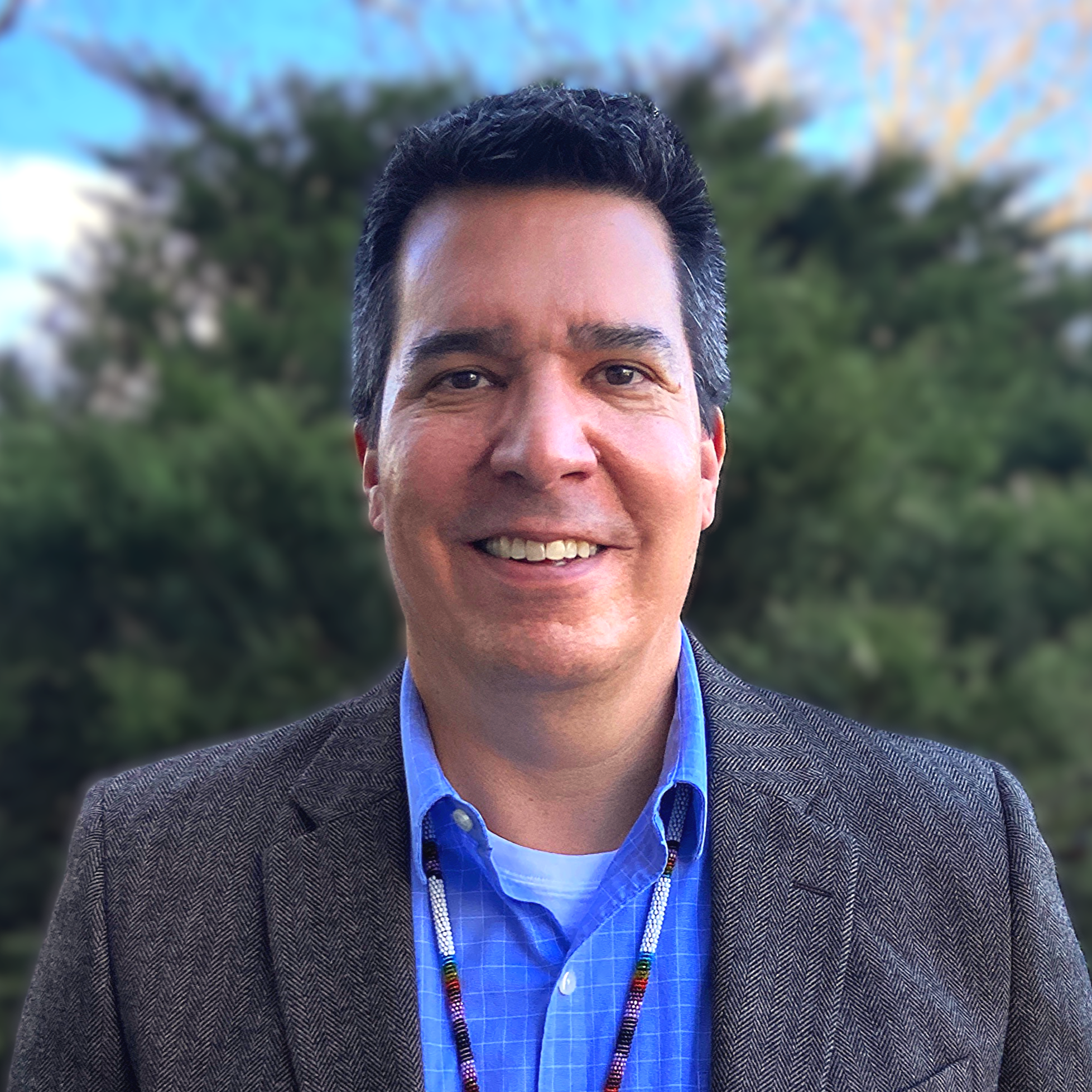 Man in blue striped button down shirt and grey suit coat smiling standing in front of green shrub background