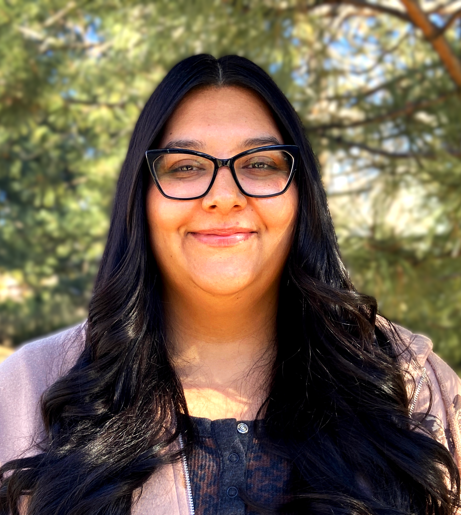 Woman with long dark hair and glasses, smiling, with a blurred background of tree branches