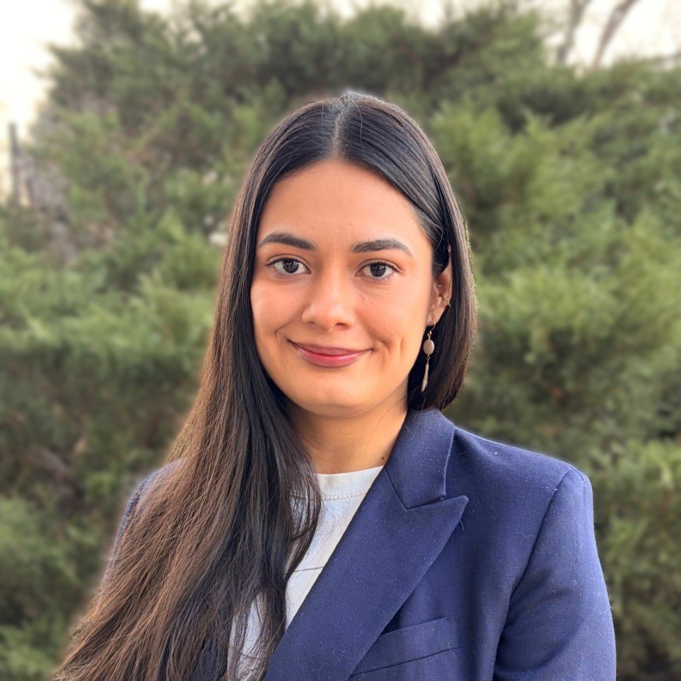 A woman with long dark hair over one shoulder wearing a blue blazer and white shirt and standing in front of a background of greenery smiles at the camera